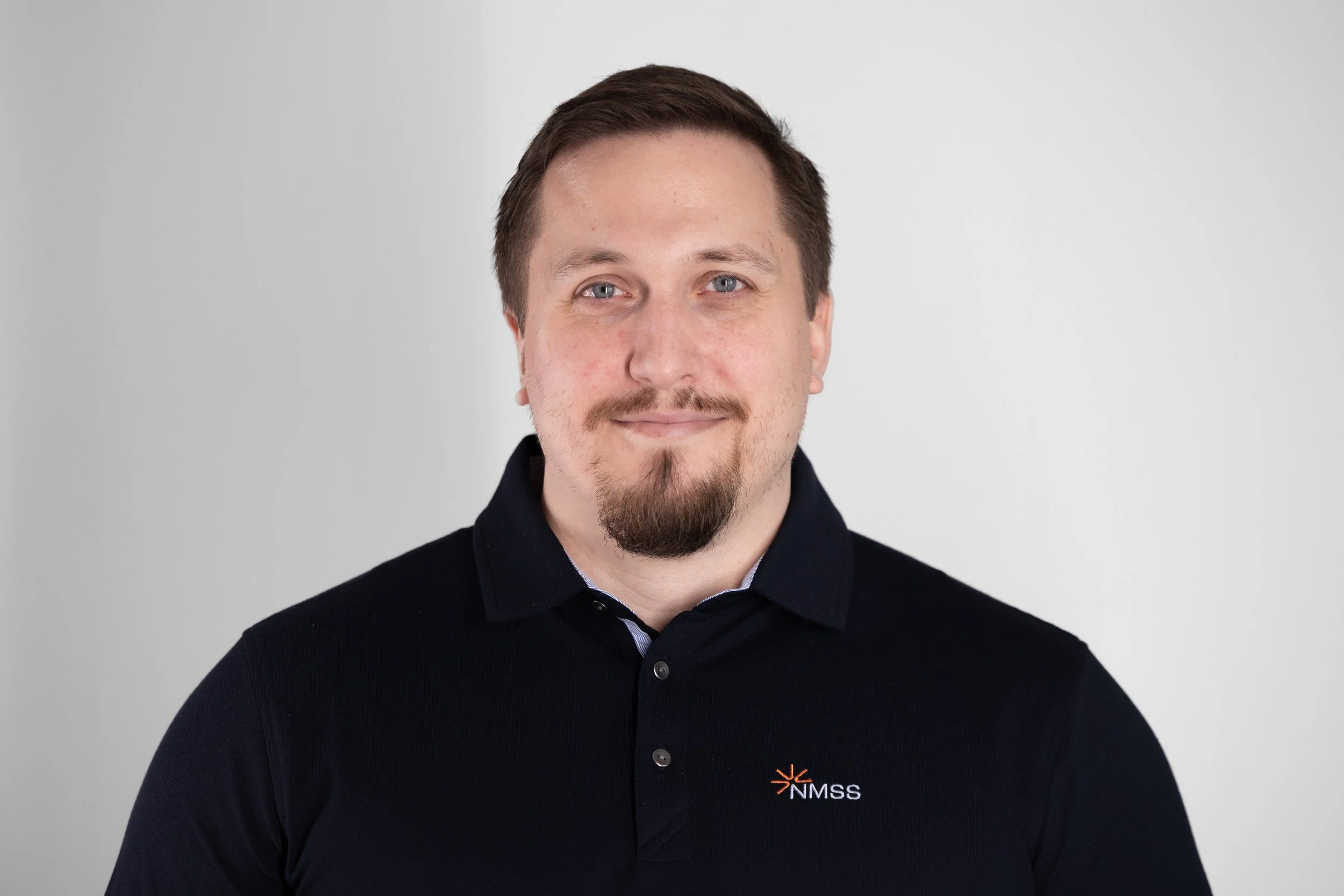 Portrettbilde av en mann med kort, mørkt hår, i en mørkeblå piquesjorte med NMSS-logo som står mot en lys bakgrunn.