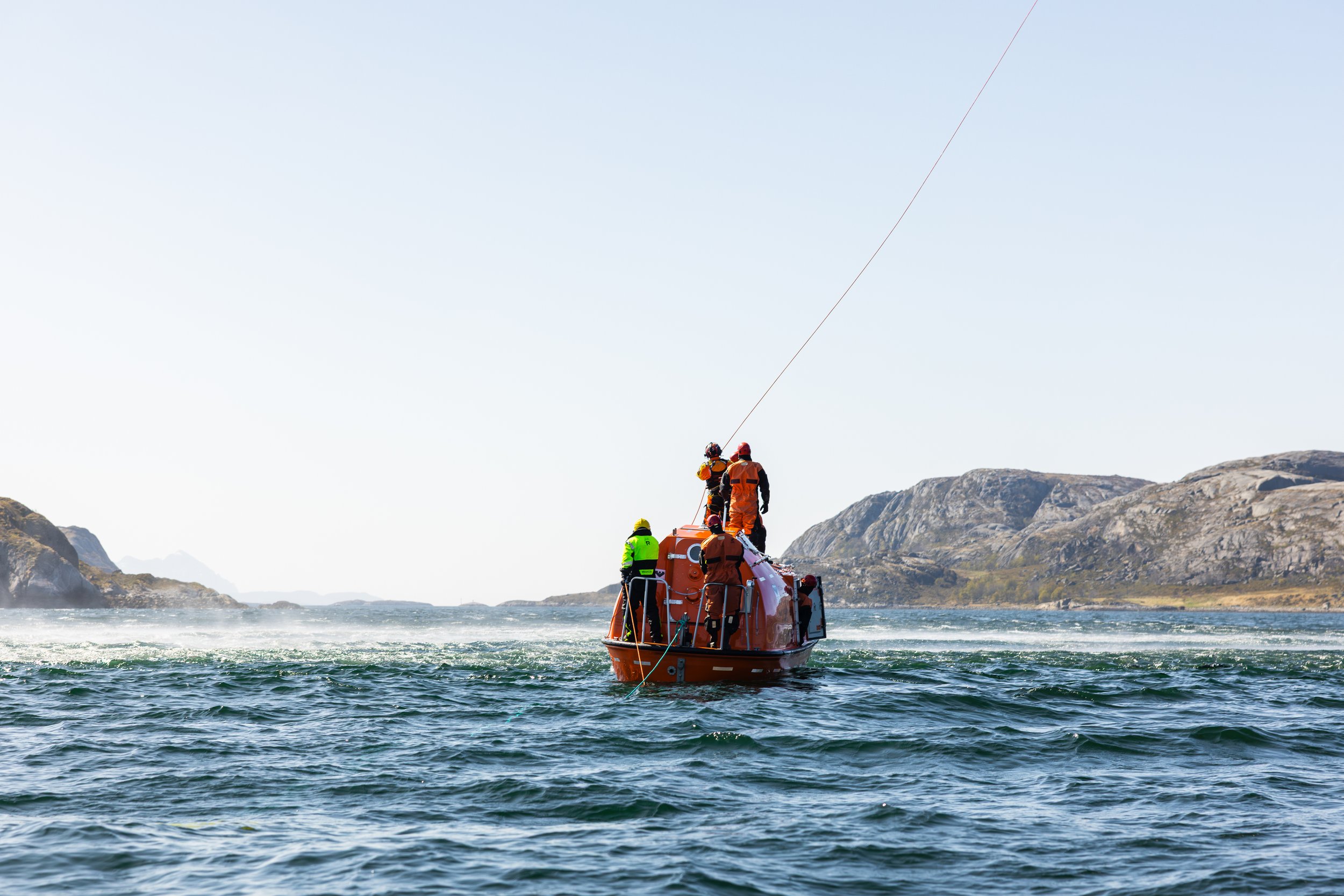 En oransje redningsbåt med fire personer i redningsdrakter om bord, i havet under en klar himmel og fjell i bakgrunnen.