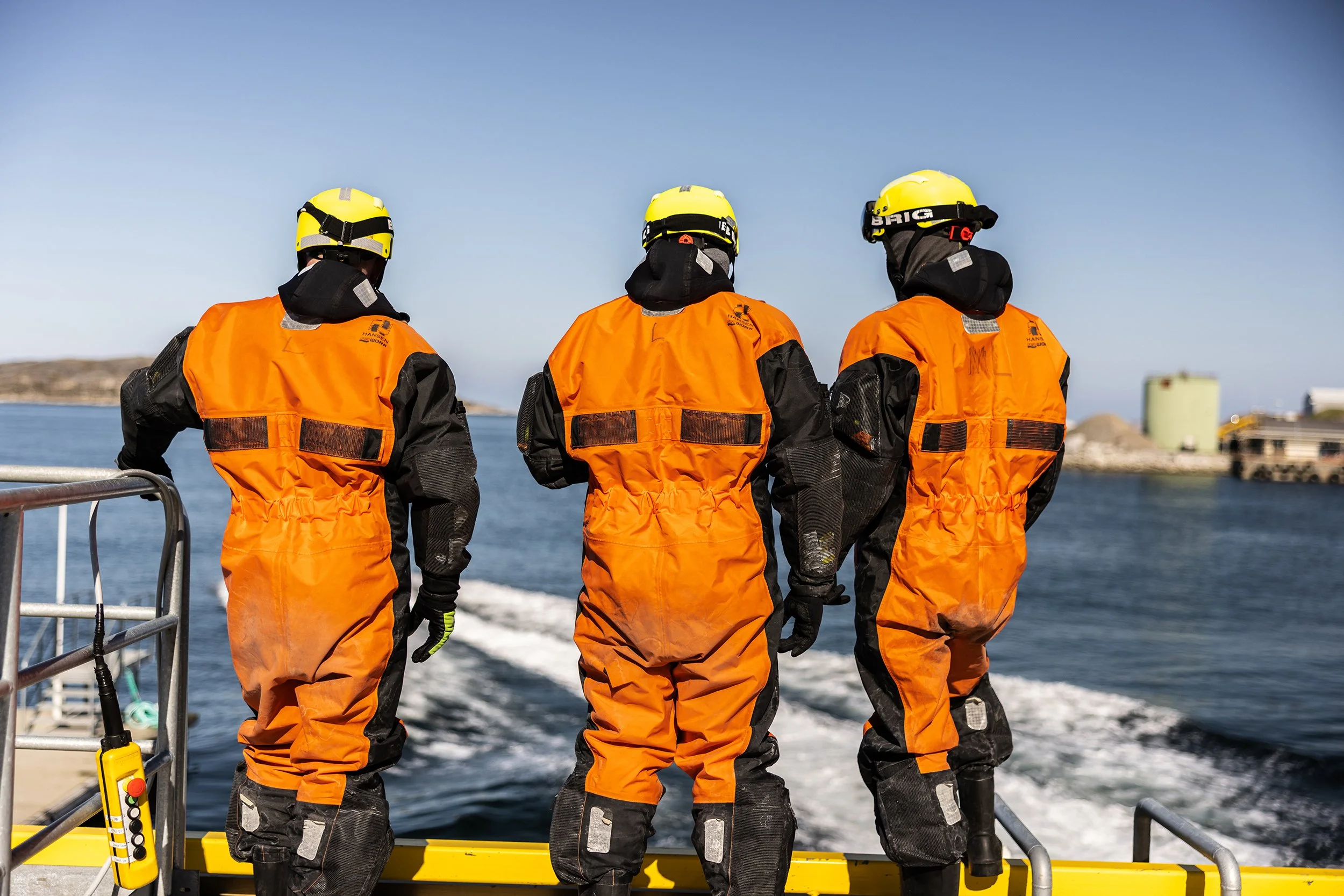 Tre personer i oransje og svarte redningsdrakter med hjelmer på står på båten og ser mot havet.