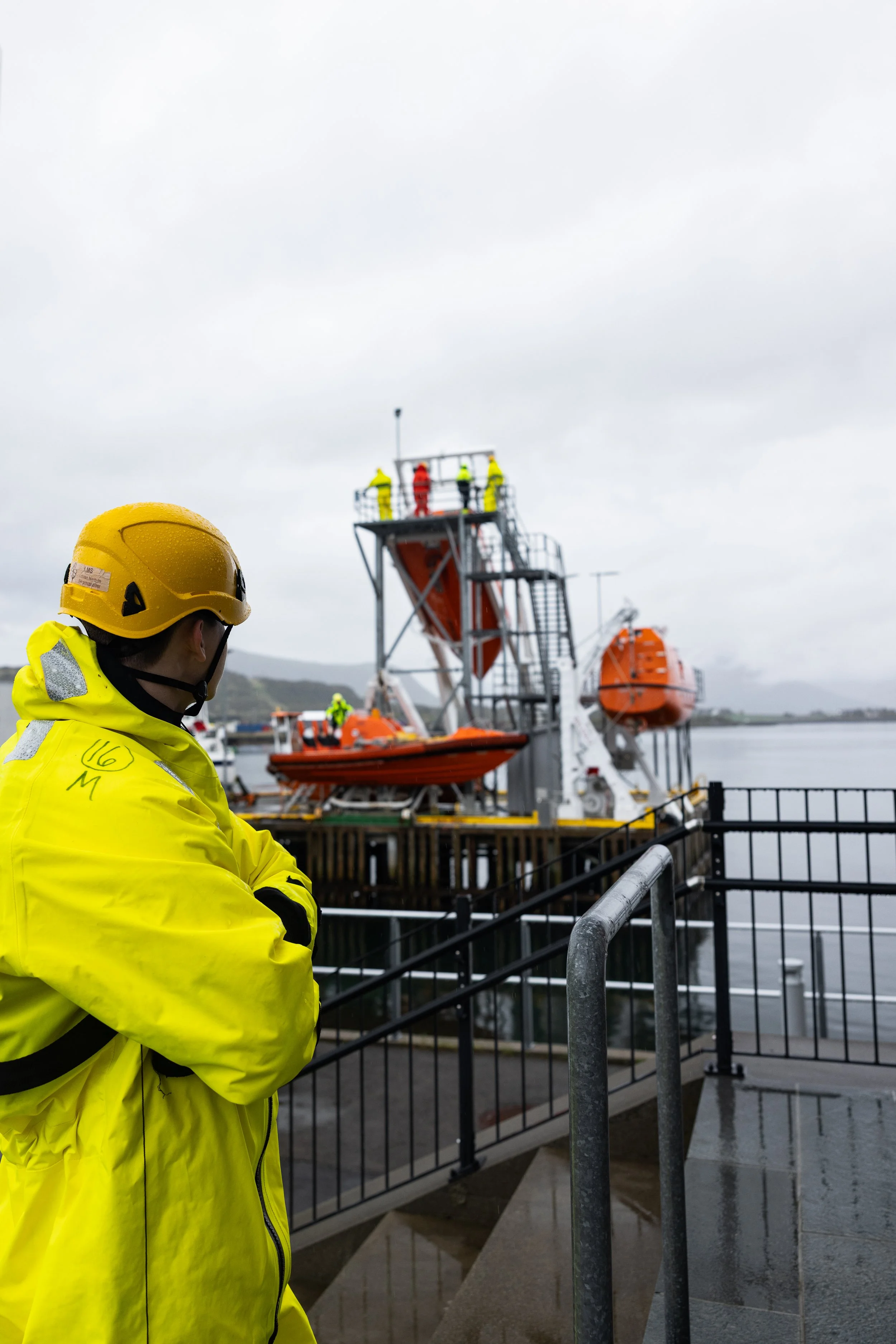En person i gul redningsdrakt og hjelm står foran en båtdavit på kaia, med flere personer i gule redningsdrakter på plattformen til daviten. Dette er en sikkerhetsøvelse ved sjøen.