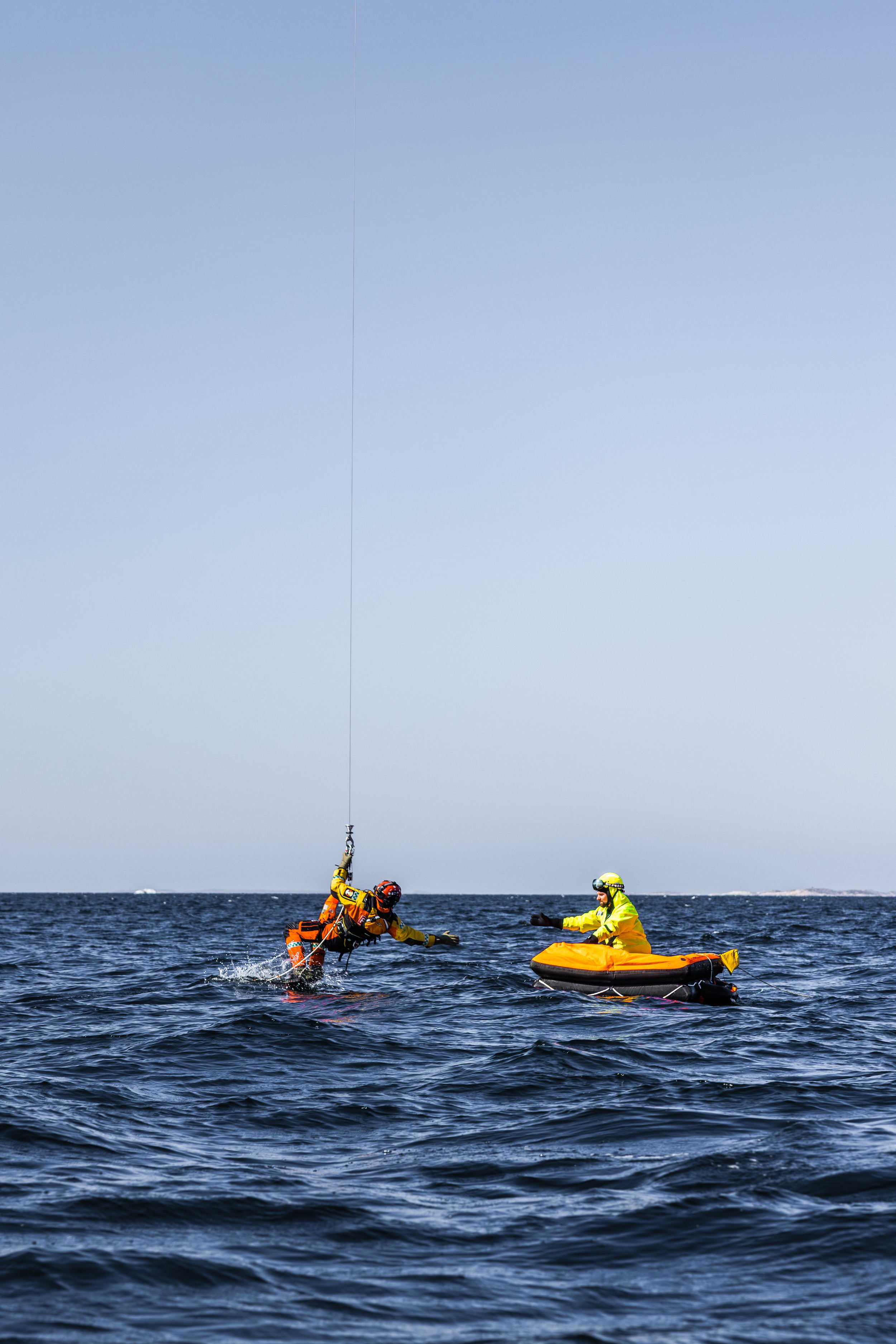 En sjøøvelse med en redder i oransje redningsdrakt henger i tau og redder en annen person i gul redningsdrakt som sitter på en redningsflåte på havet under en klar himmel.