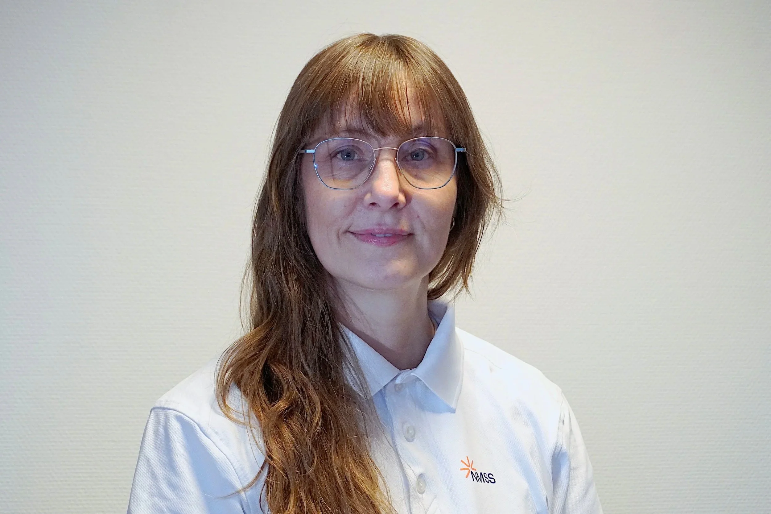 Portrettbilde av en kvinne med langt hår i en hvit piquesjorte med NMSS-logo som står mot en lys bakgrunn.