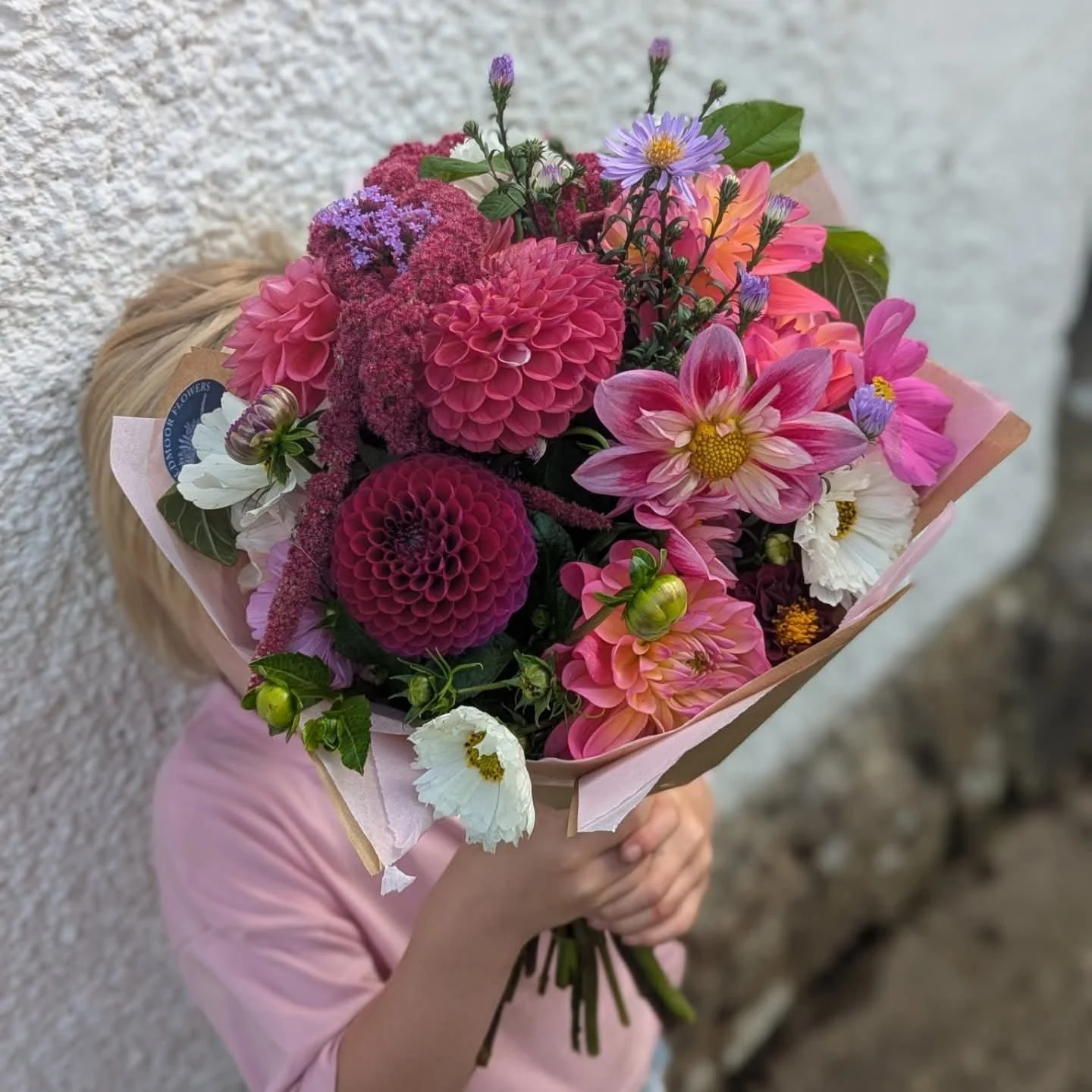 Child holding a colorful bouquet of pink, purple, white, and orange flowers standing against a textured wall.