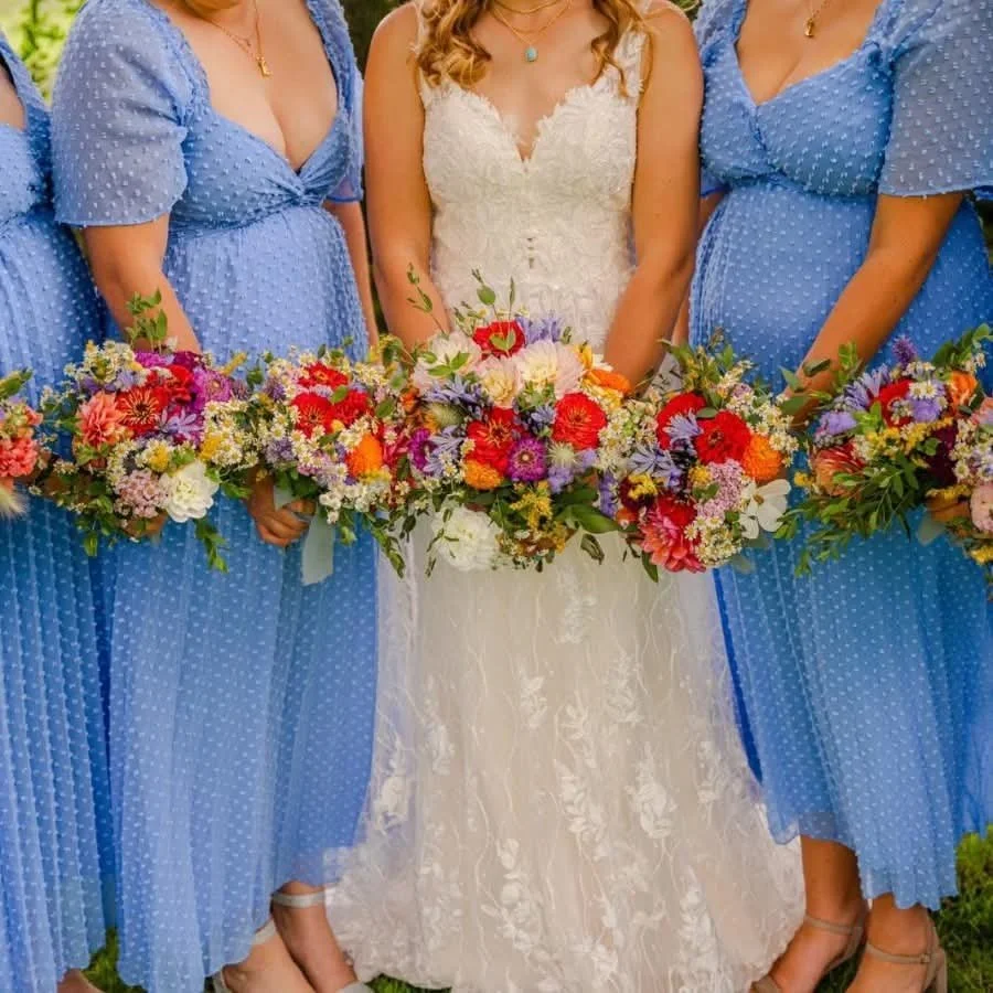 A bride in a white wedding dress standing between women in blue polka dot dresses, all holding colorful floral bouquets.