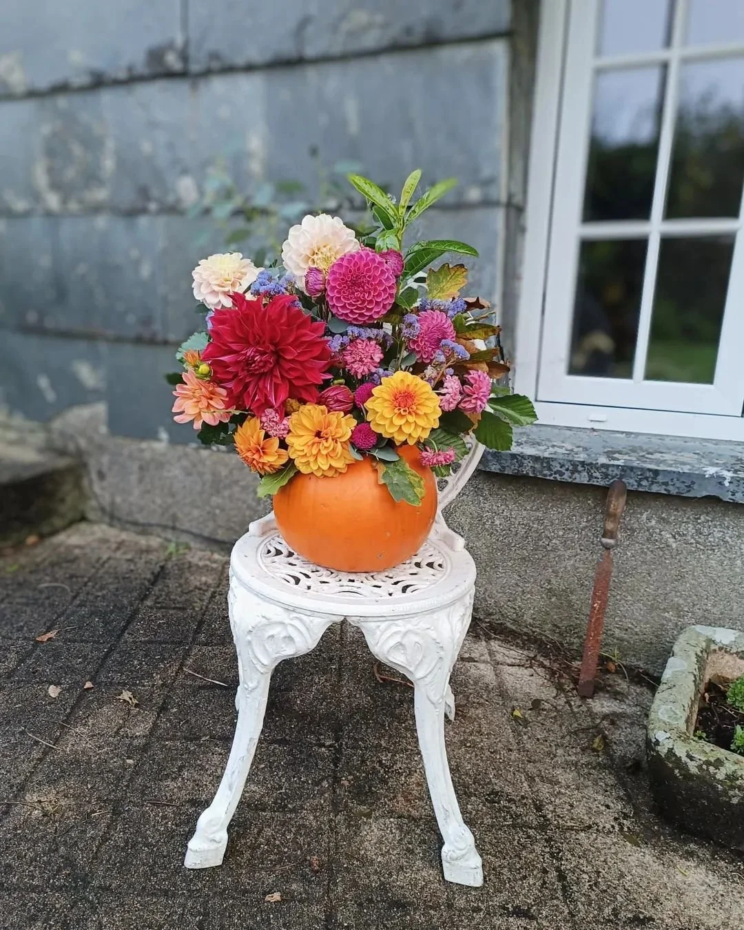 A pumpkin-shaped vase filled with colorful flowers, sitting on an ornate white stool outside near a stone wall and a window.