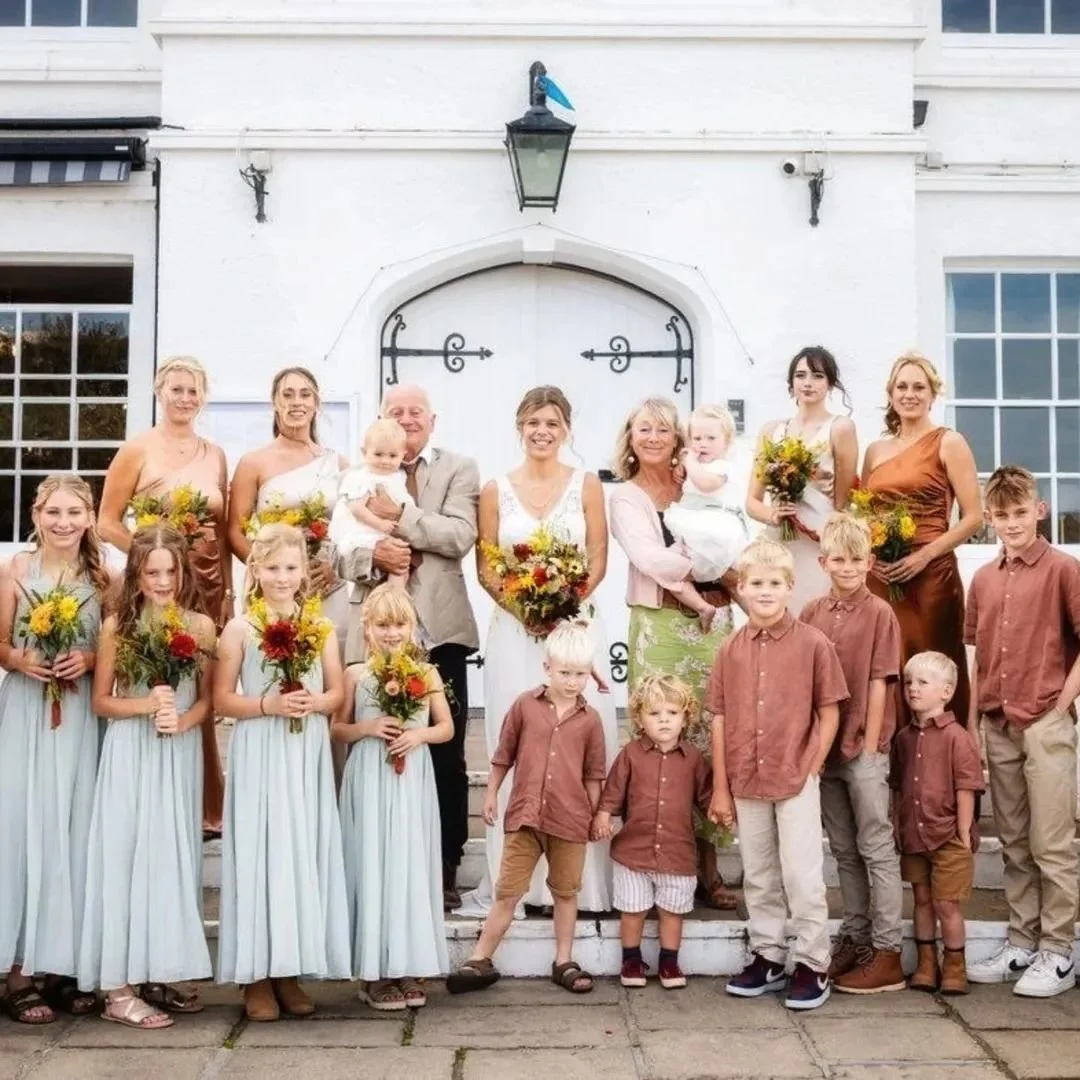 Group of people, including children and adults, standing outdoors in front of a white building with a large door, during a formal event like a wedding, with some holding colorful bouquets.
