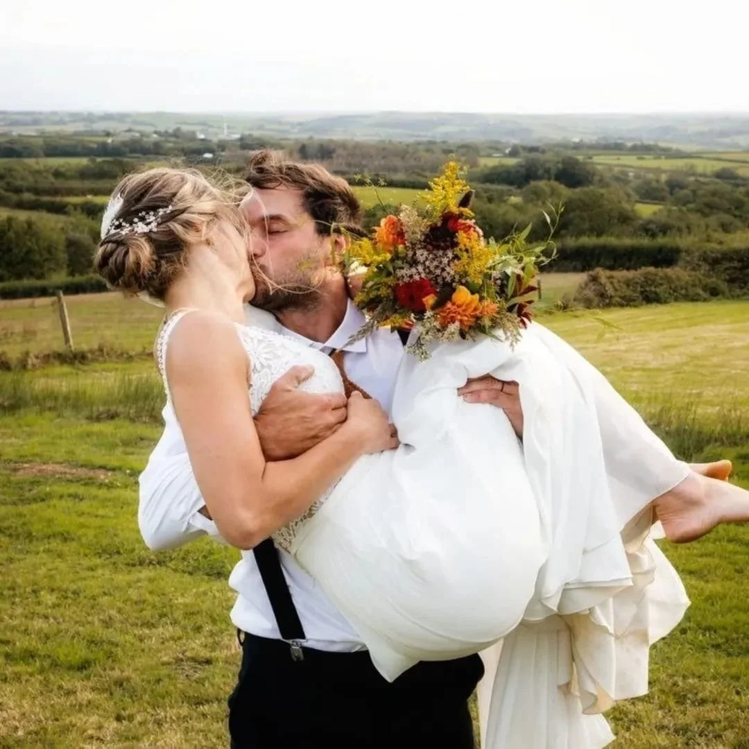 A man in a white shirt and suspenders holding a woman in a white wedding dress and holding a large bouquet of flowers, as they share a kiss outdoors on a green field with rolling hills in the background.