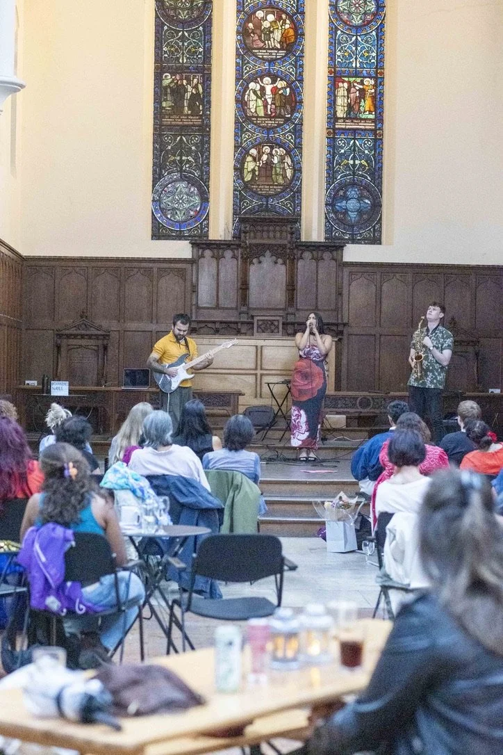 Musicians performing on stage in front of a choir and audience inside a church with stained glass windows.