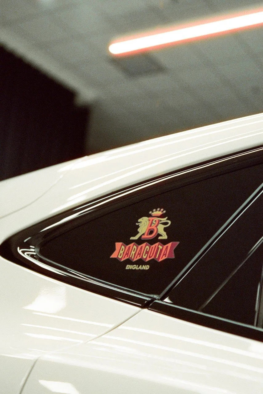 Close-up of a car window with a Baracuta England logo sticker and a partial view of the car's white body.