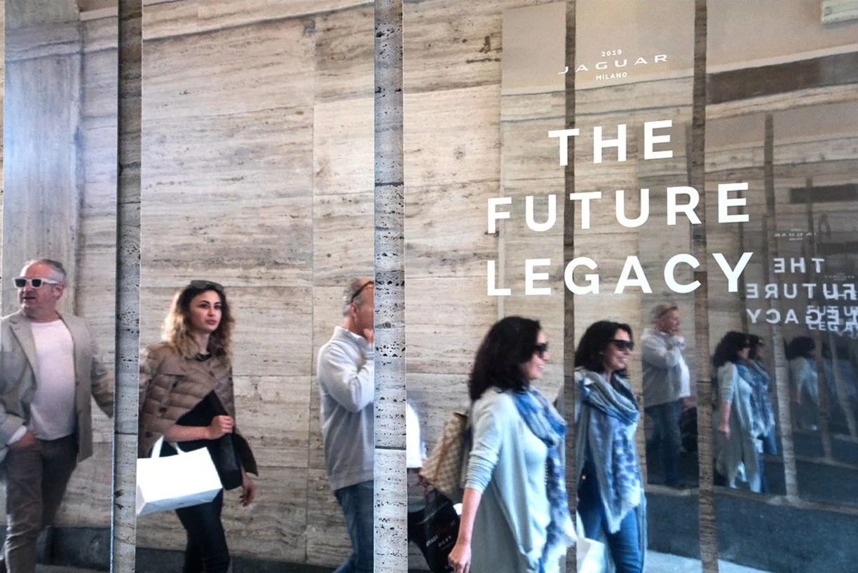 People walking past a shop window with mirrored reflections and "The Future Legacy" text, along with a Jaguar Milano logo in the background.