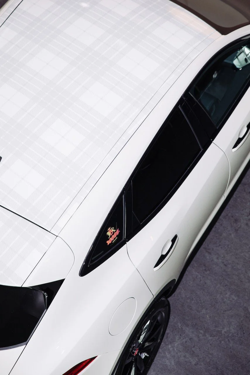 Top view of a white car with a plaid pattern on the roof and a decal on the side window, featuring dark tinted windows and a sleek design.