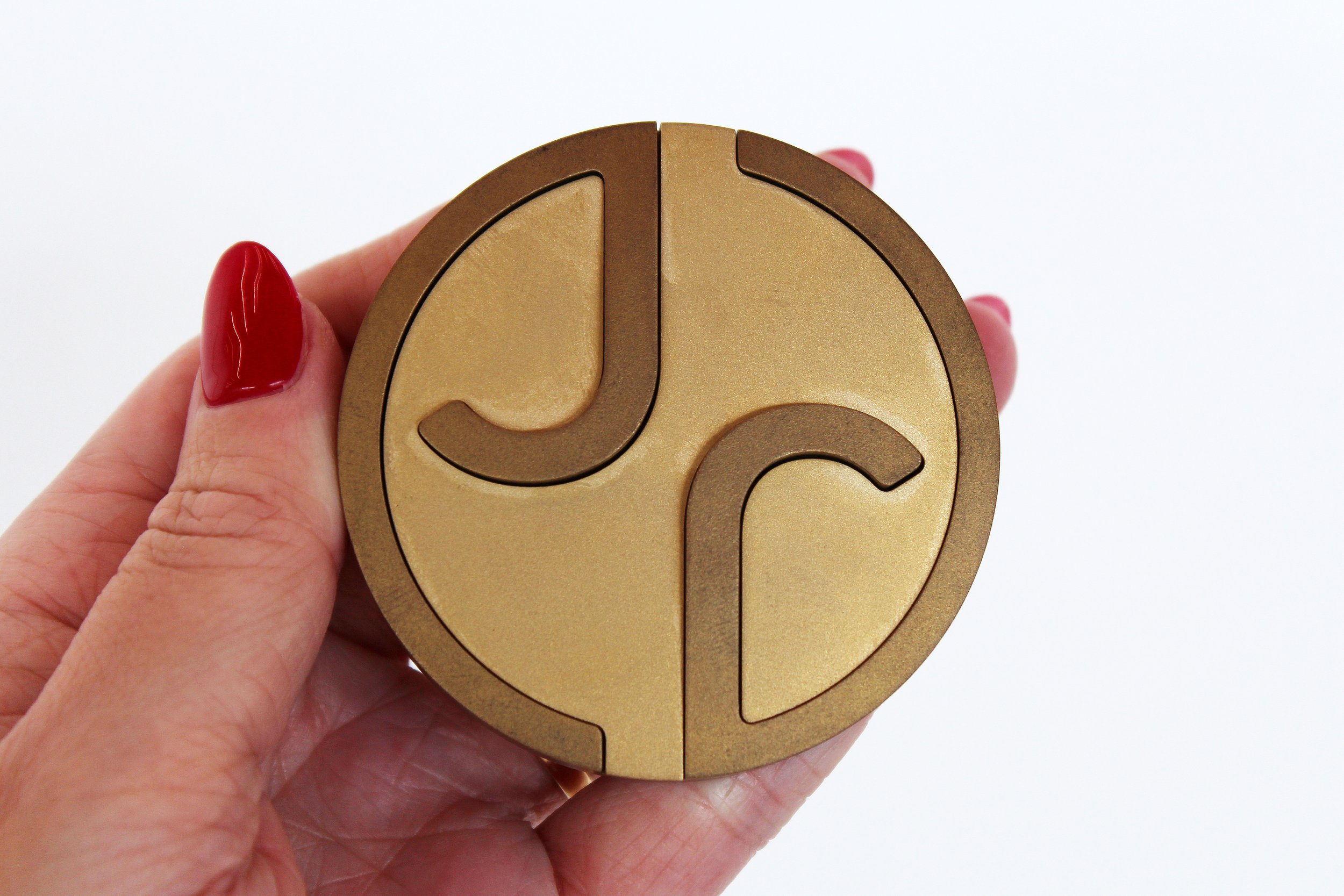A person with red nail polish holding a round, gold puzzle object with interlocking pieces on a white background.