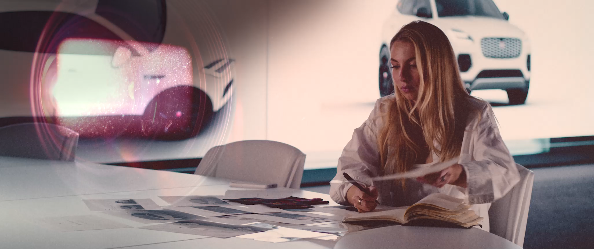Woman at a desk reviewing car design sketches with a white car in the background.