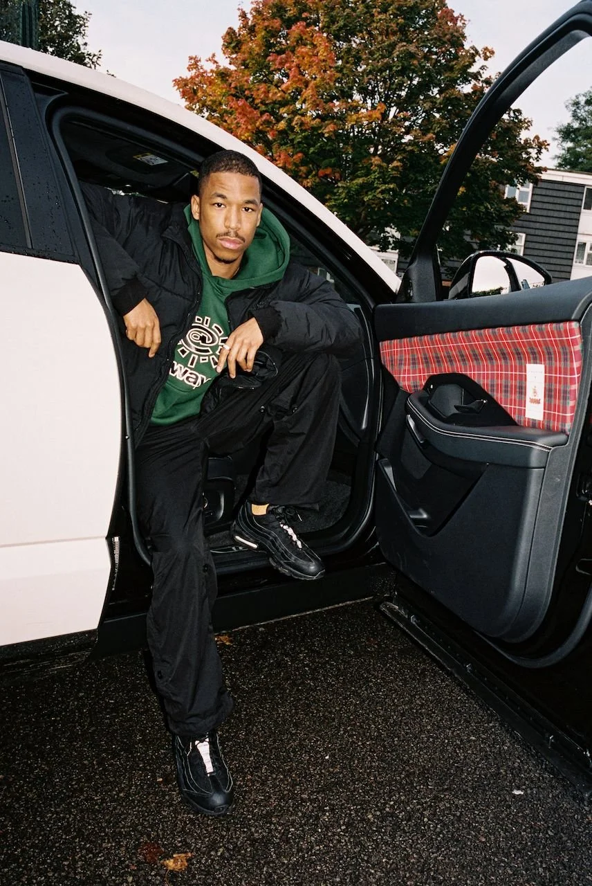 Person sitting on the edge of an open car door wearing a black jacket, green hoodie, and black sneakers. The car door features a plaid pattern. In the background, there is a tree with autumn leaves.
