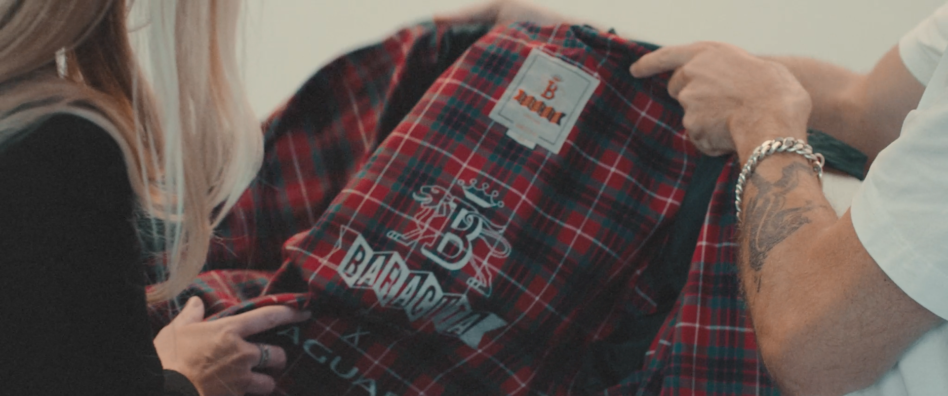 Two people holding a red plaid jacket featuring embroidered graphics and logos including "B Baracuta" and "Jaguar."