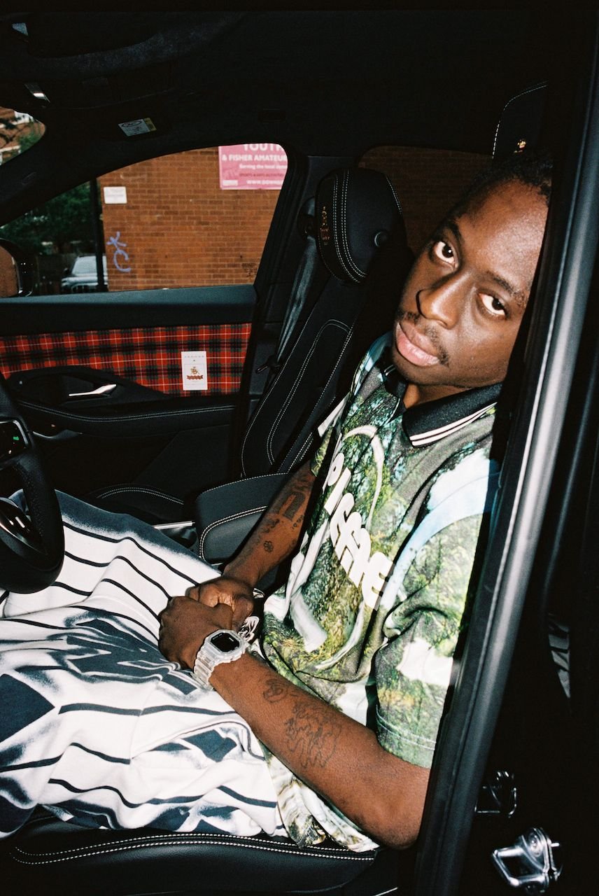 Man sitting in a car with plaid interior, wearing a graphic t-shirt and striped pants.