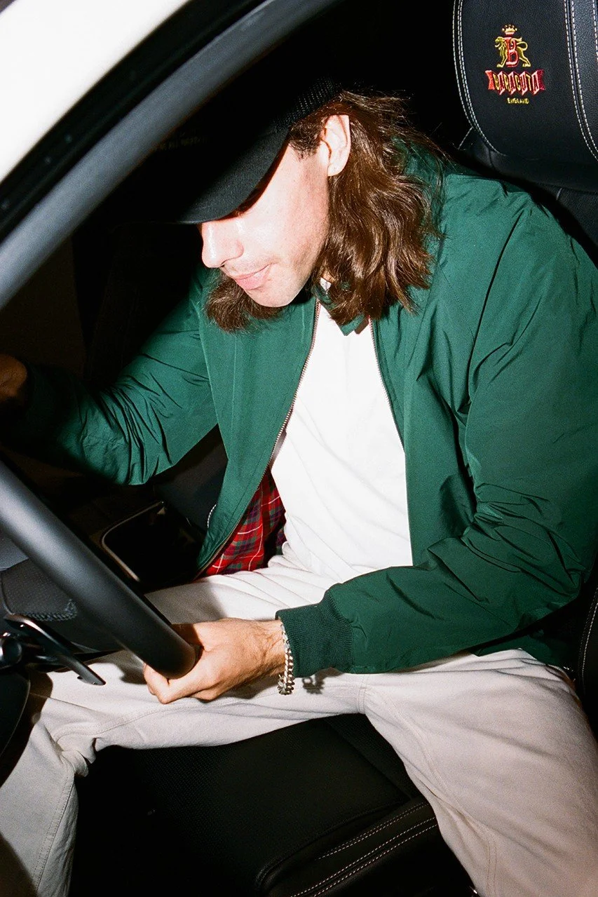 Man wearing green jacket, white shirt, and cap sitting in a car, holding the steering wheel.