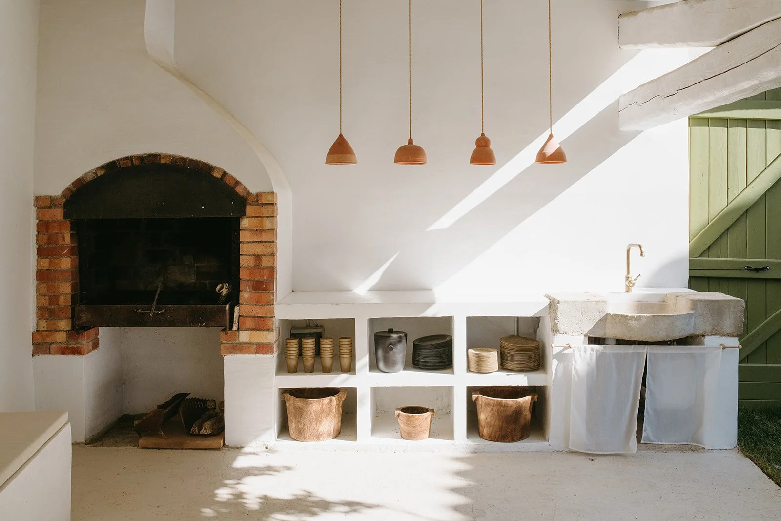 Rustic outdoor kitchen with brick fireplace, terracotta pendant lights, concrete sink, open shelving, and green wooden door
