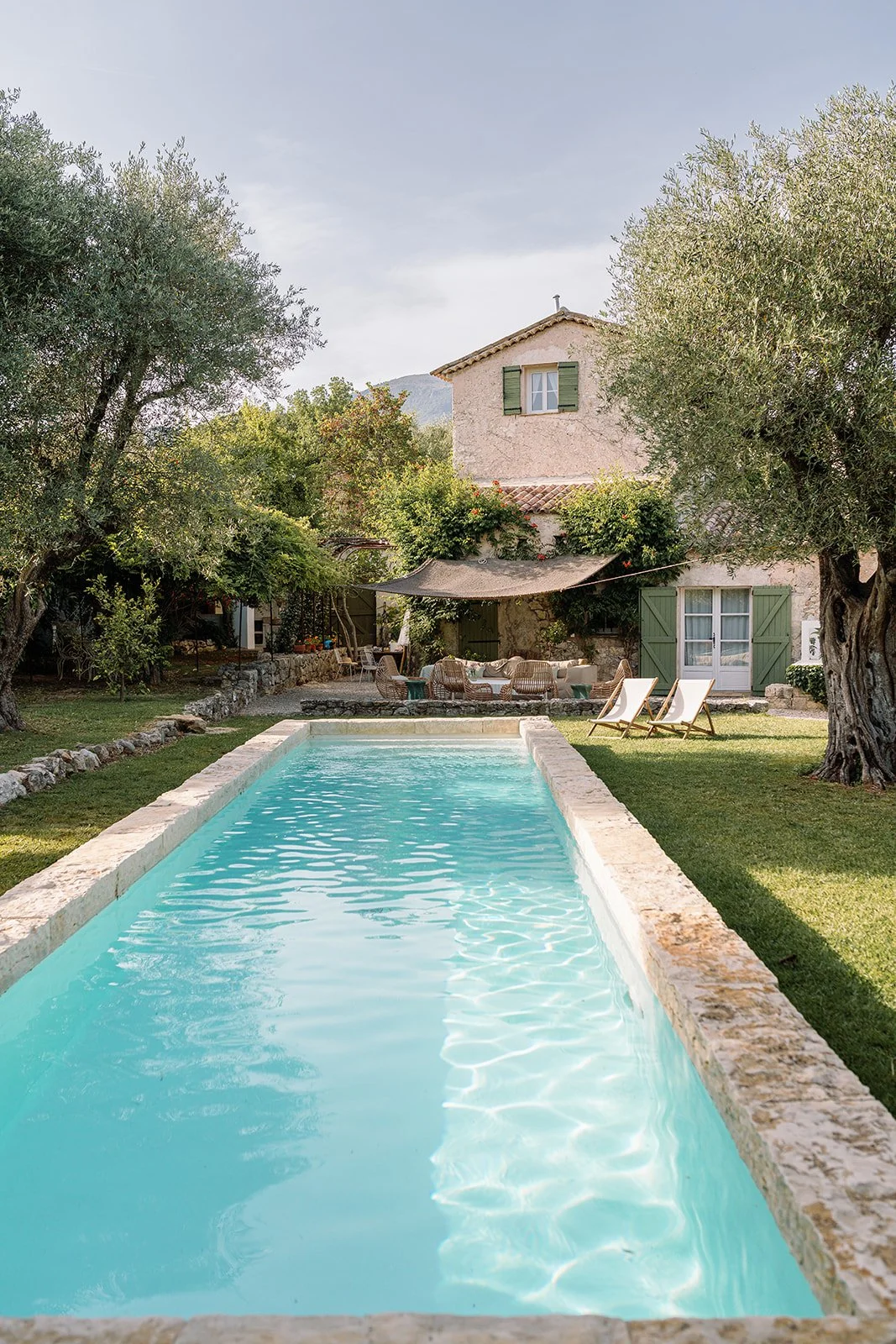 Stone farmhouse courtyard with a turquoise swimming pool, lounge chairs, patio seating, and olive trees