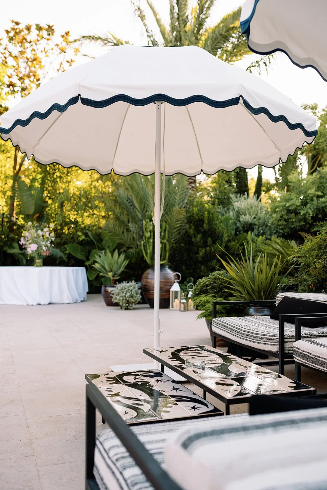 Outdoor patio with a white scalloped umbrella, patterned coffee tables, lounge chairs, and lush tropical garden