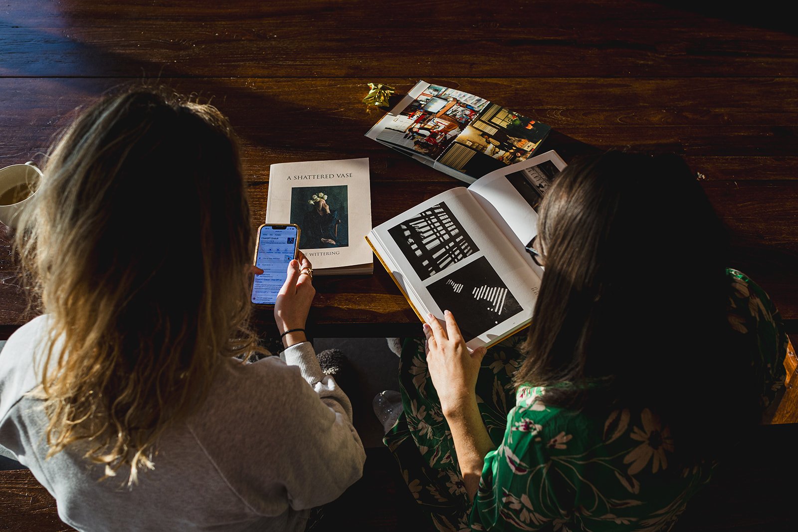 Deux femmes assises à une table en bois regardant des livres photo et un téléphone, avec des papiers et une tasse de café sur la table