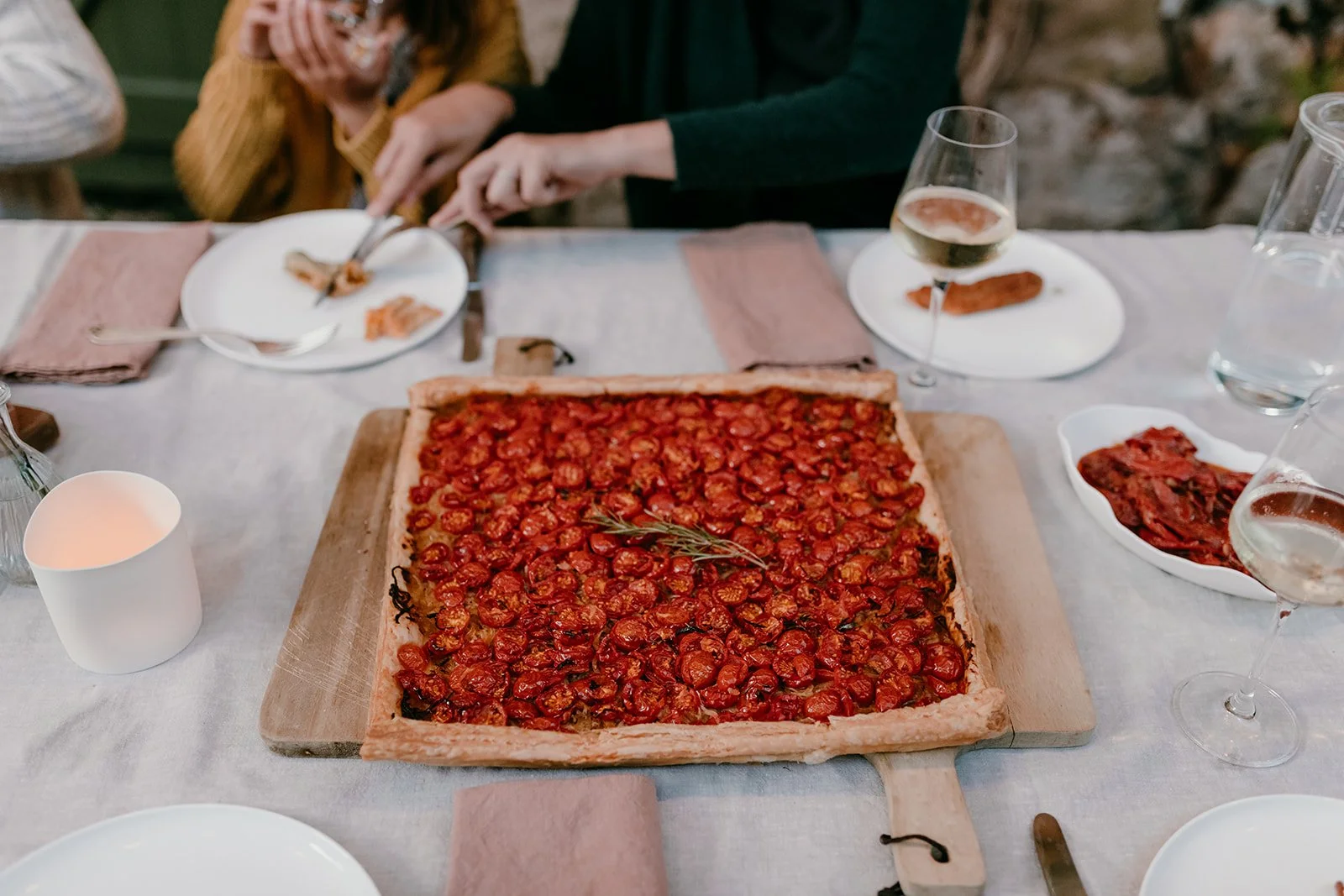 Outdoor table set for a meal with a large roasted tomato tart, wine glasses, plates, and diners serving food