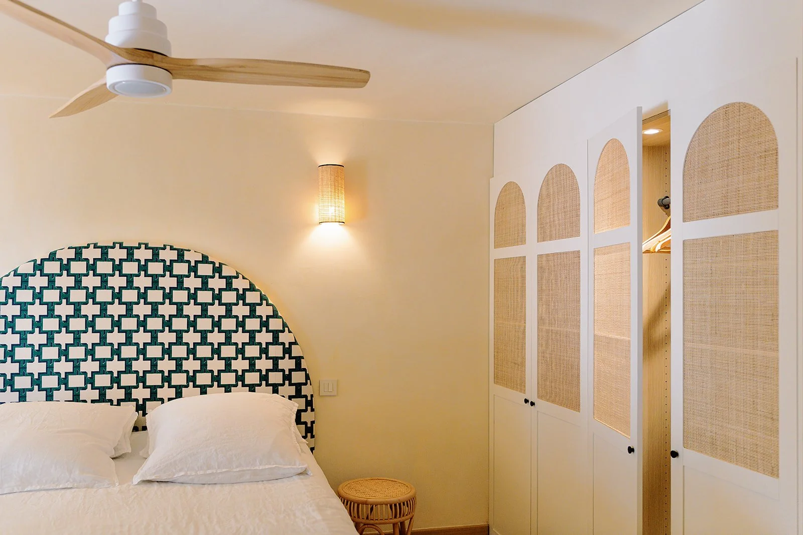 Cozy bedroom with patterned upholstered headboard, white bedding, rattan-front wardrobe, ceiling fan, and wall sconce