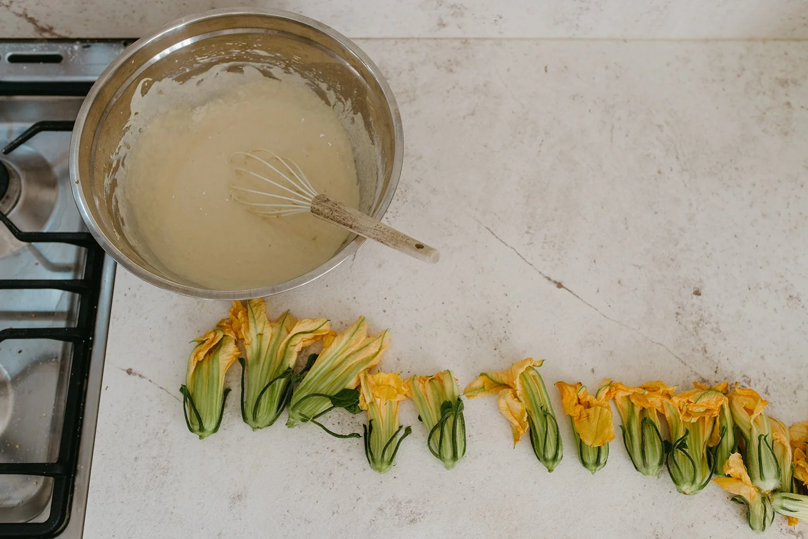 Overhead view of a bowl of creamy batter with a whisk beside a row of yellow squash blossoms on a kitchen counter.