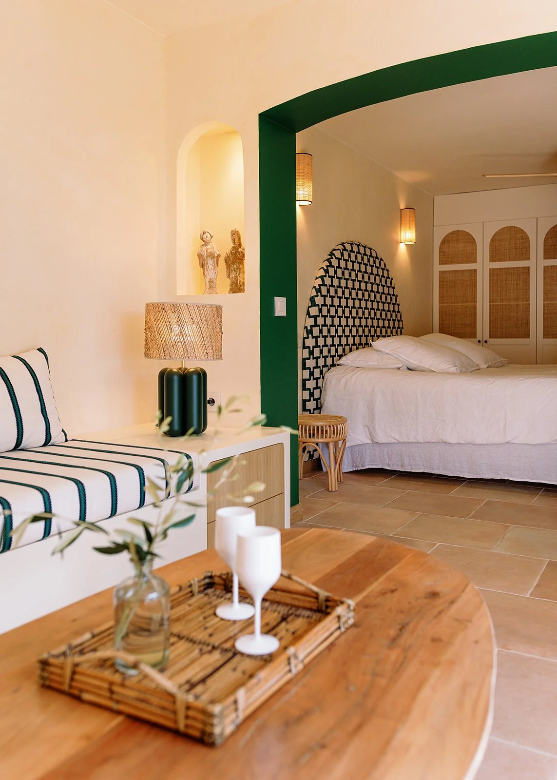 Warmly lit Mediterranean-style bedroom and sitting area with green archway, patterned headboard, rattan lighting, striped bench, and wooden table with tray
