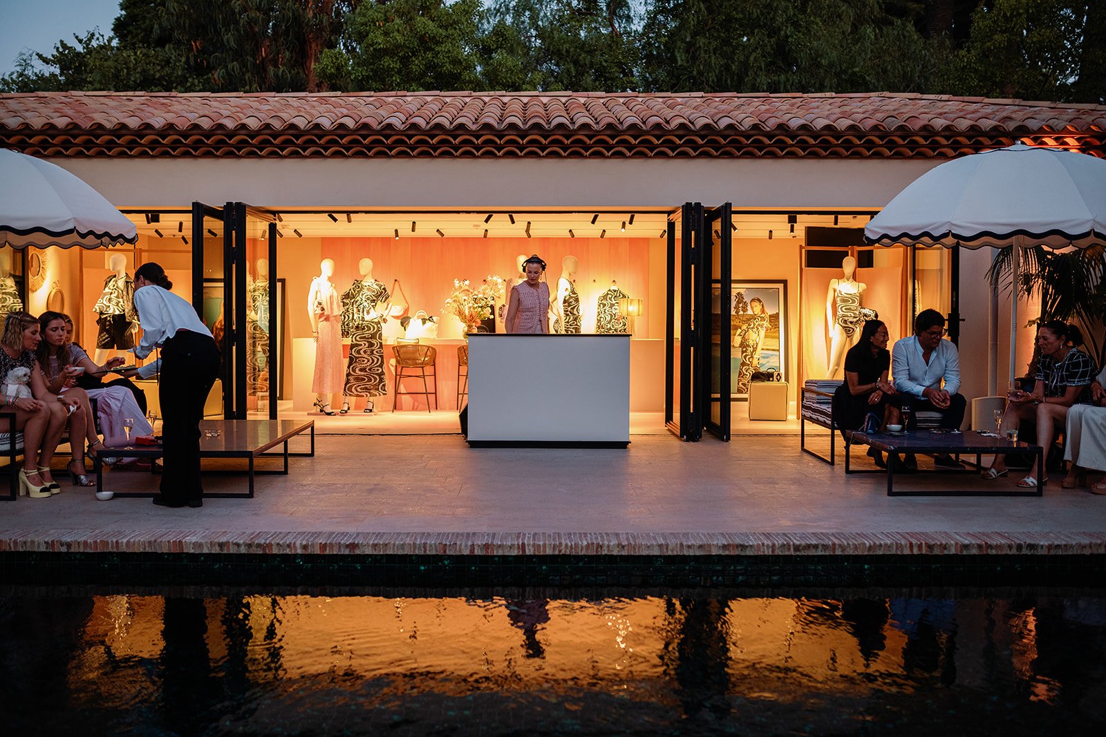 Outdoor fashion boutique event with mannequins, guests, and a clothing display overlooking a reflective pool