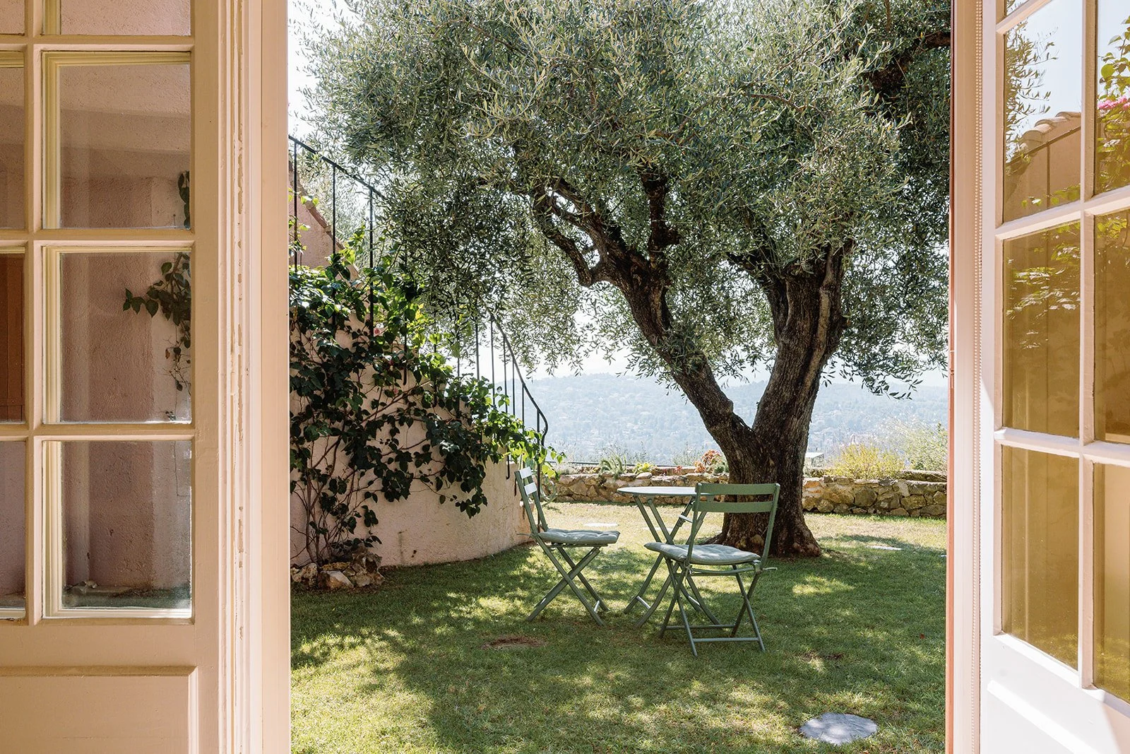 Garden patio viewed through open French doors, with two folding chairs beneath an olive tree and hills in the background