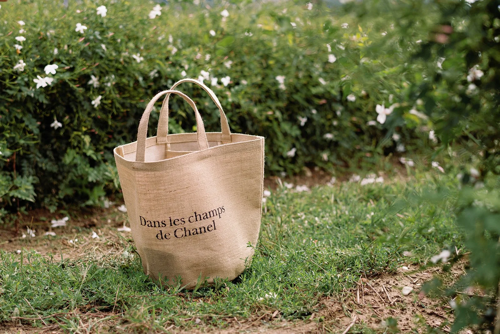 Natural jute tote bag reading “Dans les champs de Chanel” resting on grass in a flowering garden
