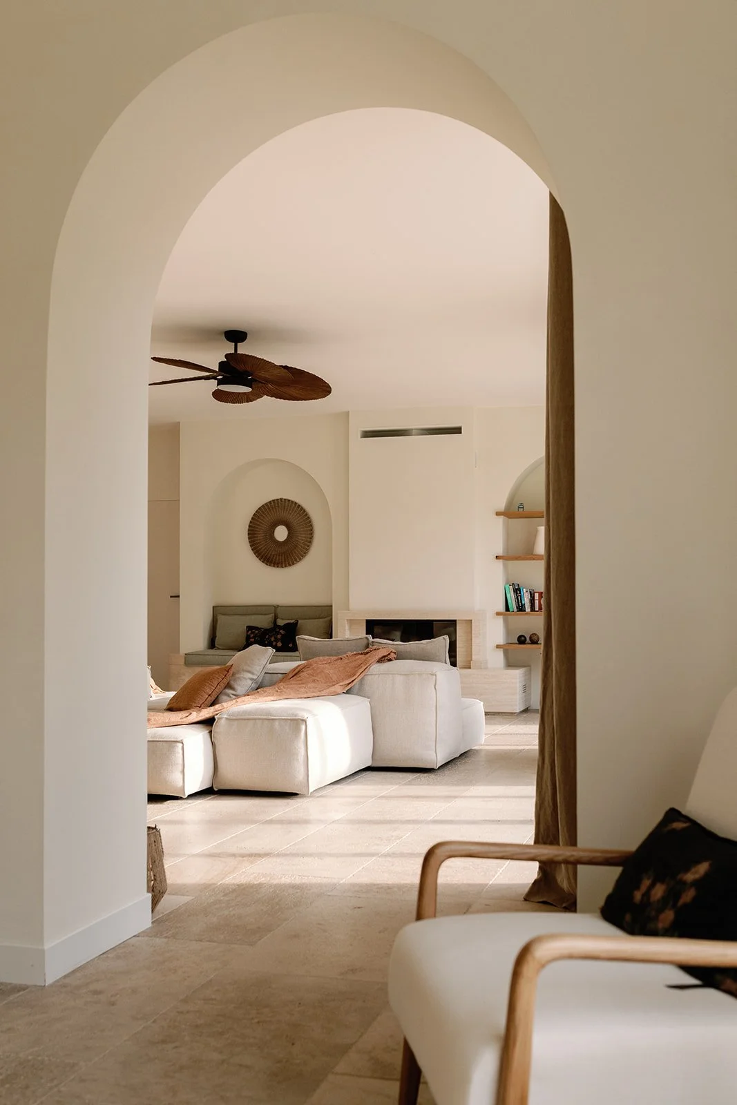 Warm minimalist living room viewed through an arched doorway, featuring a ceiling fan, white sectional sofa, fireplace, built-in shelves, and woven wall decor.