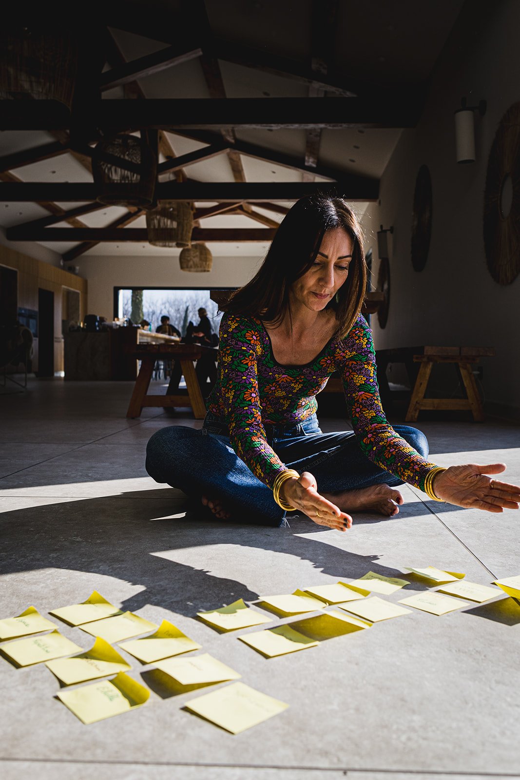 Une femme assise au sol, jambes croisées, disposant des post-it par terre dans une pièce aux poutres en bois et au plafond haut, éclairée par la lumière naturelle.