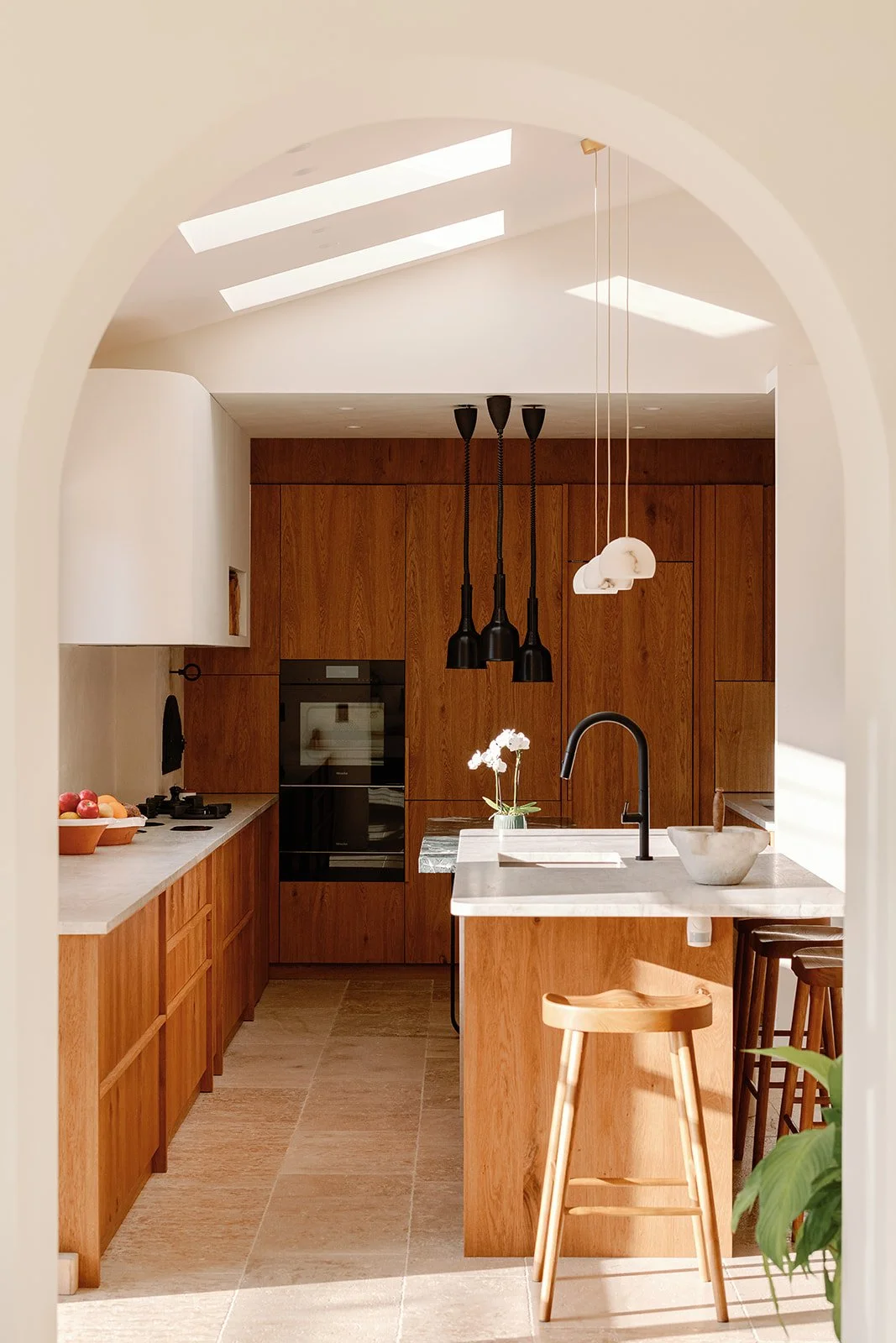Warm modern kitchen with walnut cabinetry, marble island, black pendant lights, arched doorway, and wooden barstools