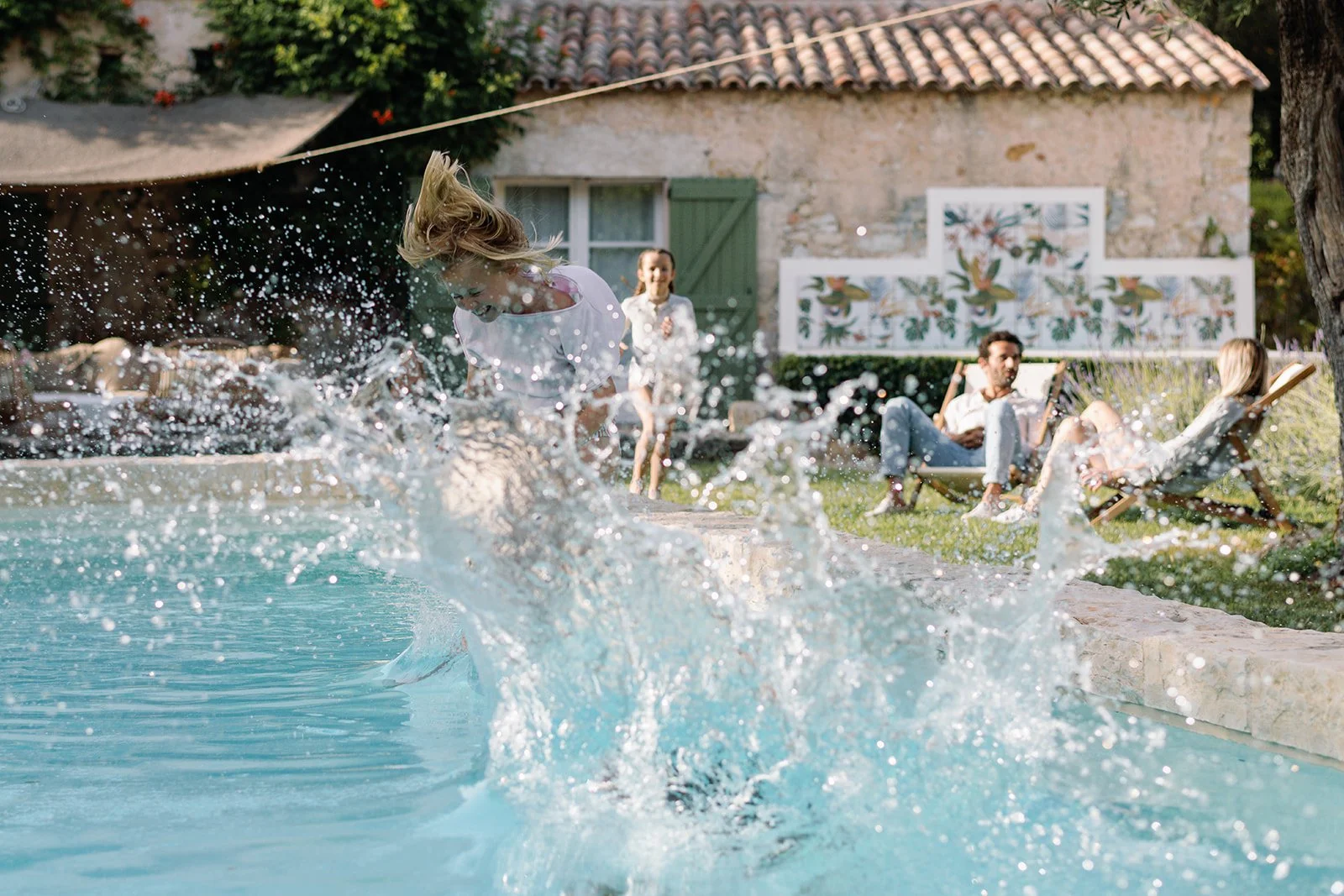Person splashing into a swimming pool while friends relax on lounge chairs at a rustic countryside house