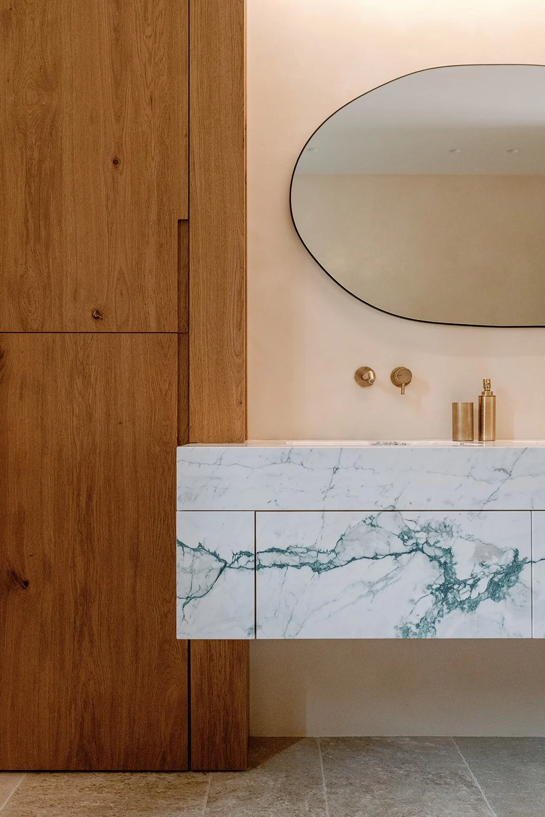 Modern bathroom vanity with a marble floating countertop, brass fixtures, oval mirror, and wood-paneled wall