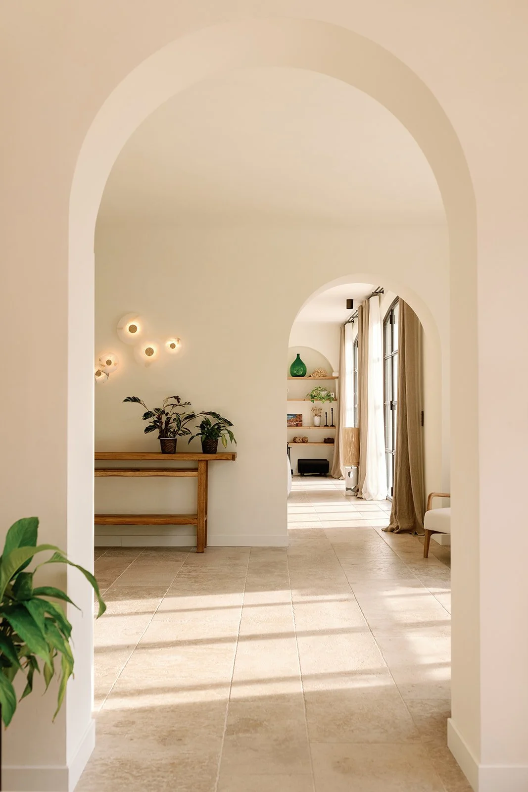 Sunlit Mediterranean-style interior with arched doorways, potted plants, wooden console table, wall sconces, shelves, and tall curtained windows