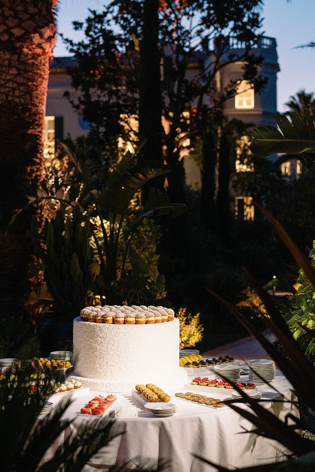 Elegant outdoor dessert table featuring a large white cake topped with powdered pastries, macarons, cookies, and fruit at dusk.