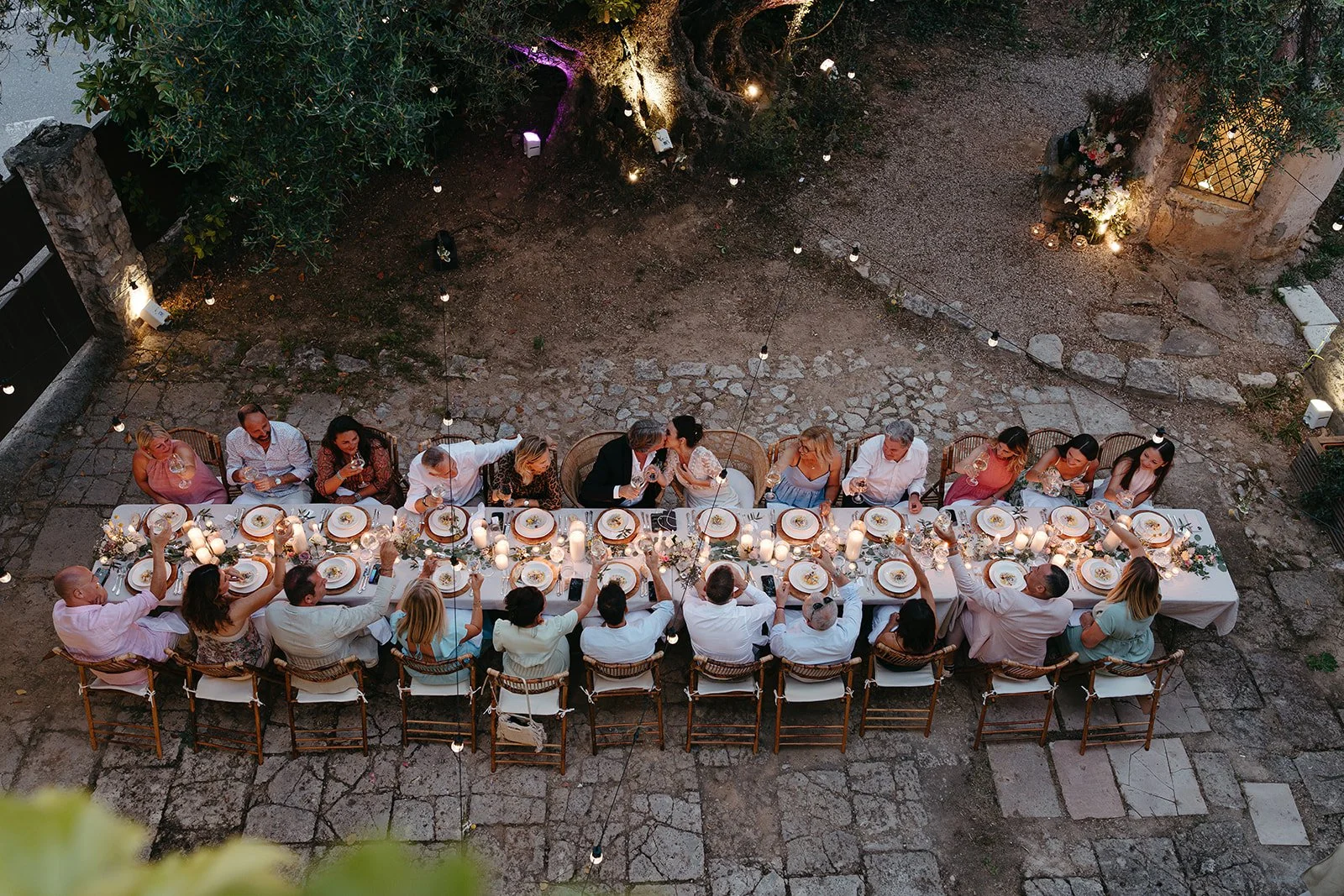 An overhead view of a long dining table outdoors at dusk, with guests raising glasses in a toast. The table is decorated with candles and floral arrangements.