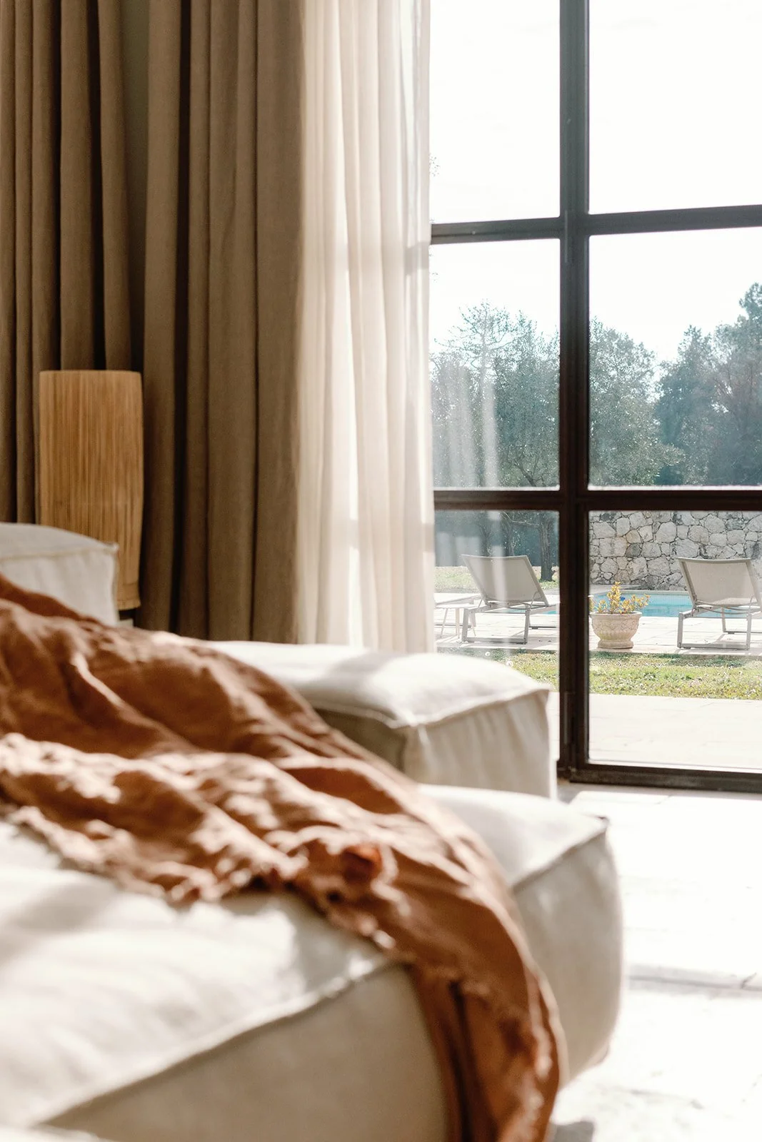 Sunlit bedroom with brown drapes, white bed, and view of a pool and lounge chairs through a large window