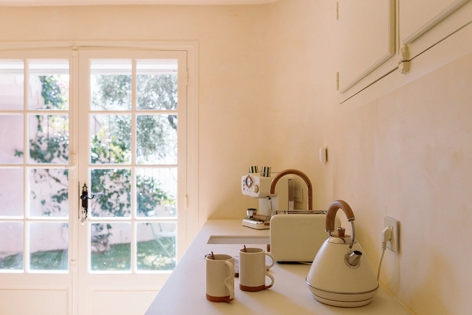 Bright kitchen counter with two mugs, a white electric kettle, toaster, and glass-paneled door