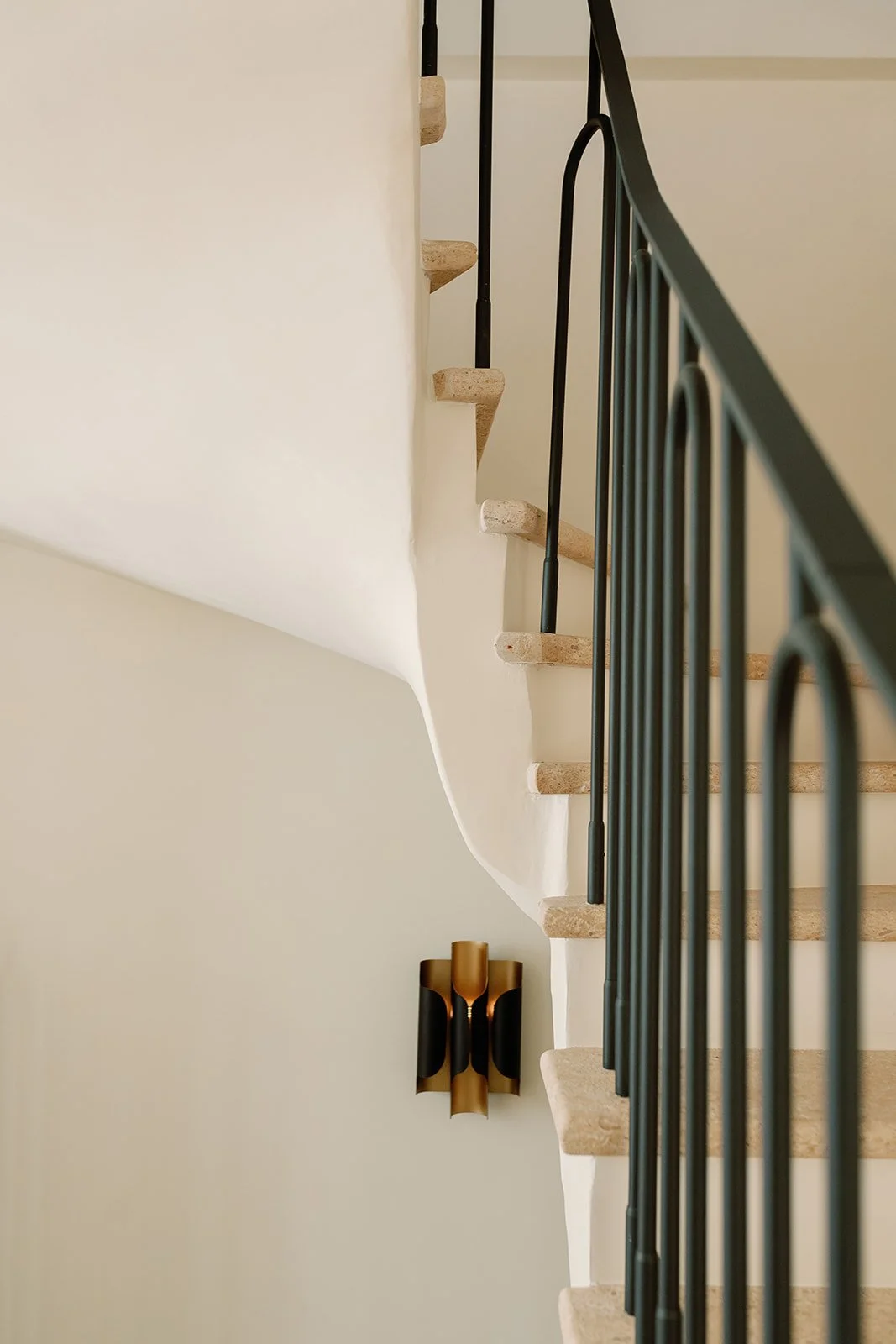 Curved staircase with cream stone steps, black metal railing, and a modern gold-and-black wall sconce.