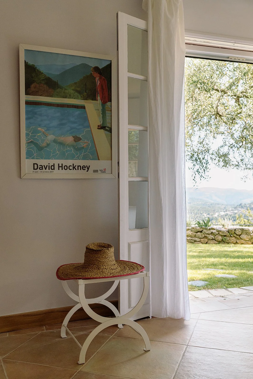 Framed David Hockney pool painting beside an open glass door, straw hat on a white table, and sunny mountain garden view.