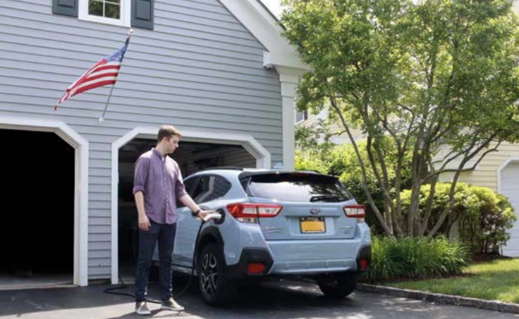 A young man is charging a blue car parked in a driveway outside a house with light blue siding. An American flag is hanging on the house, and there is a tree and greenery in the background.