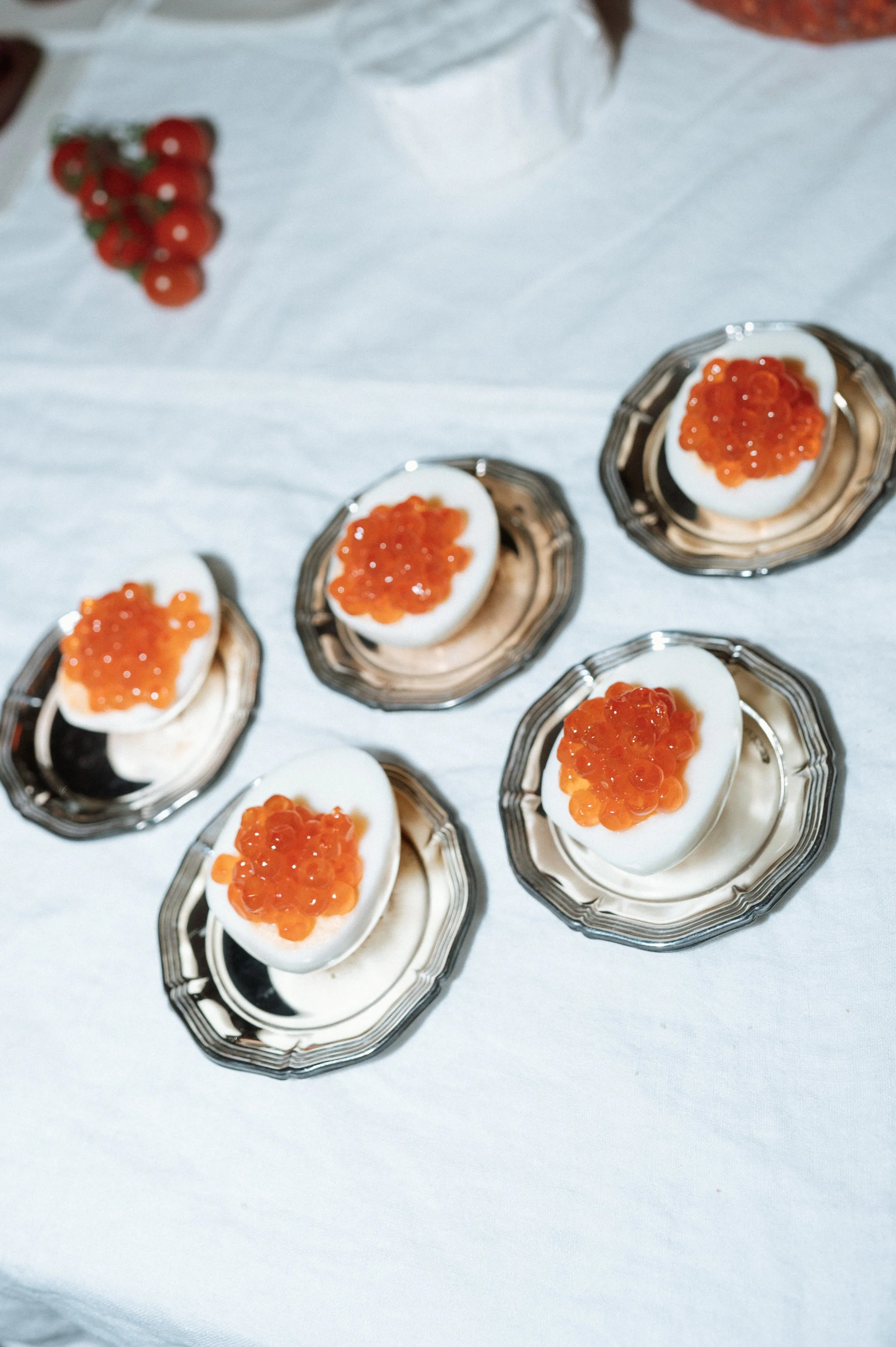 Six silver serving spoons each holding a halved boiled egg topped with orange fish roe on a white tablecloth.