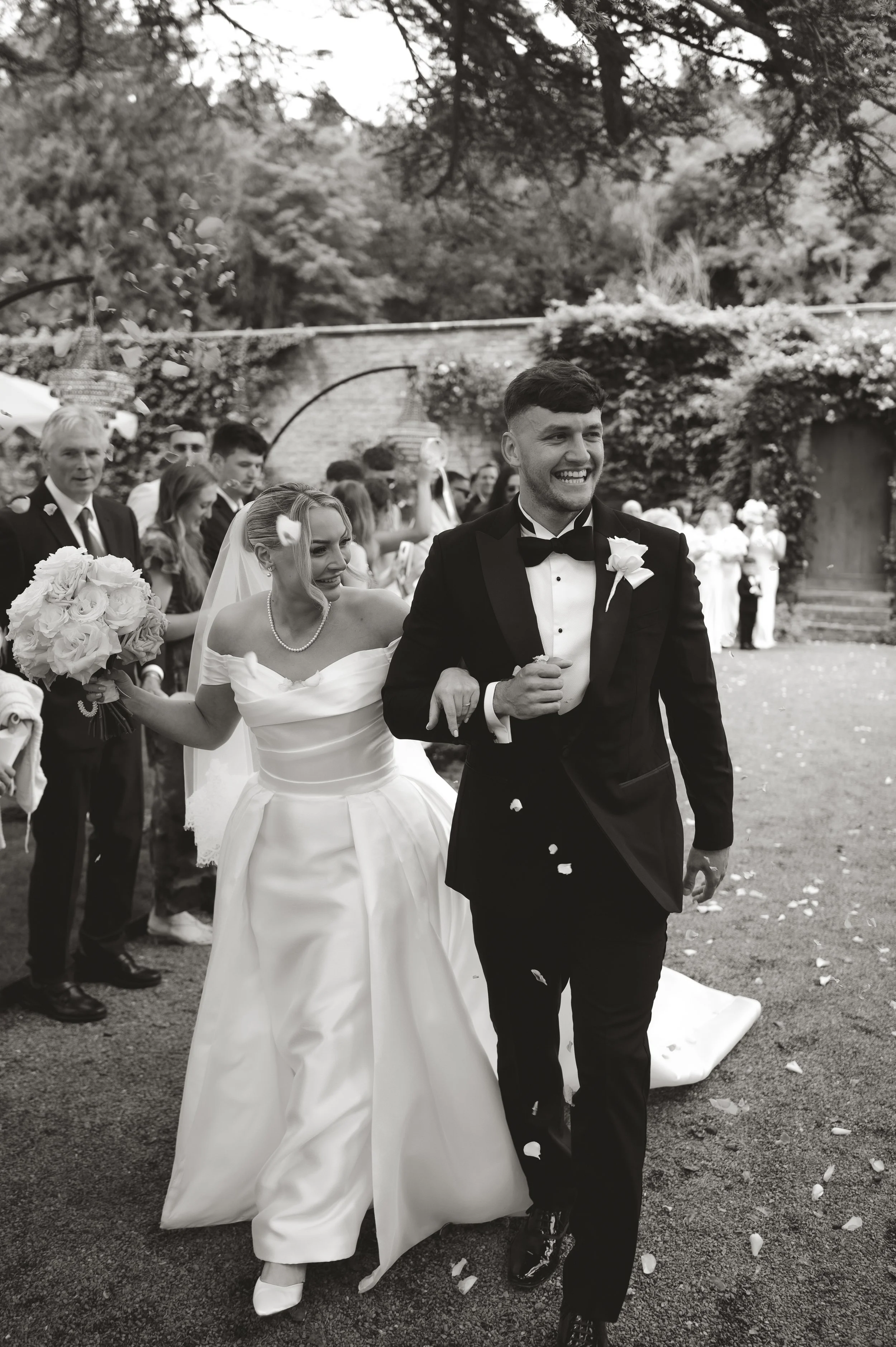 A black-and-white photograph of a wedding procession outdoors. The bride and groom are smiling, with the groom dressed in a tuxedo and the bride in a wedding gown, holding a bouquet. Guests surround them, some throwing flower petals. The background f