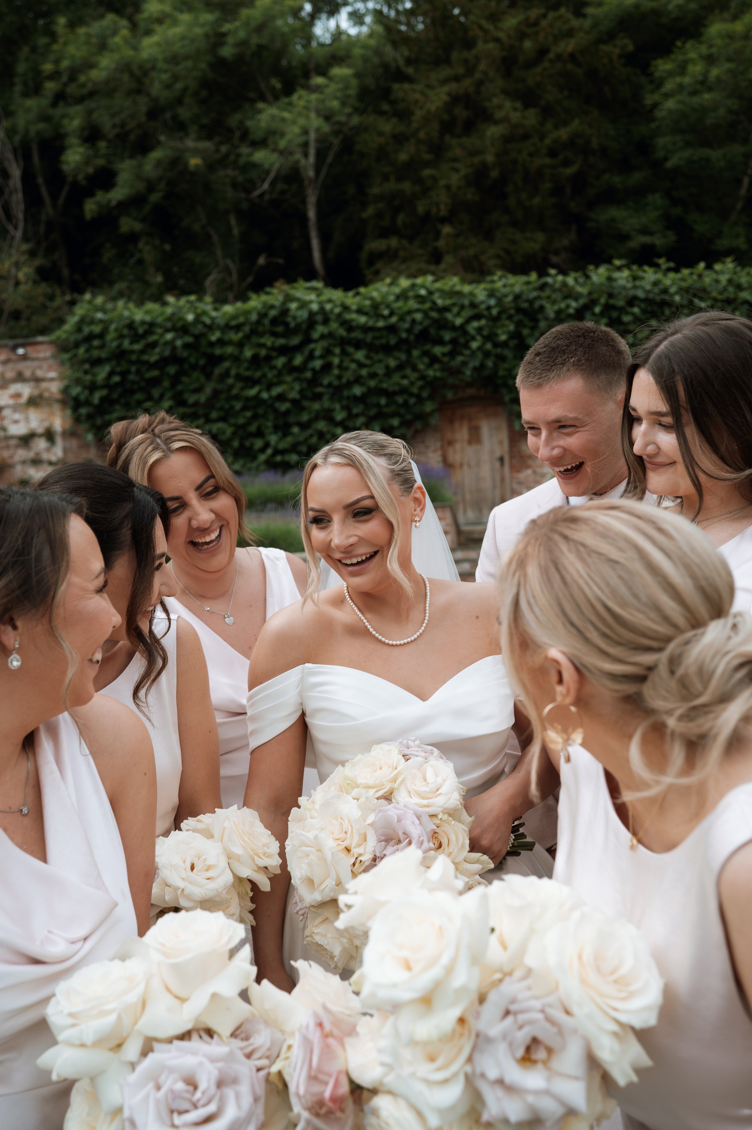 Bride in white wedding dress surrounded by bridesmaids and groomsmen, all smiling and holding bouquets of white roses, outdoors in a garden setting.