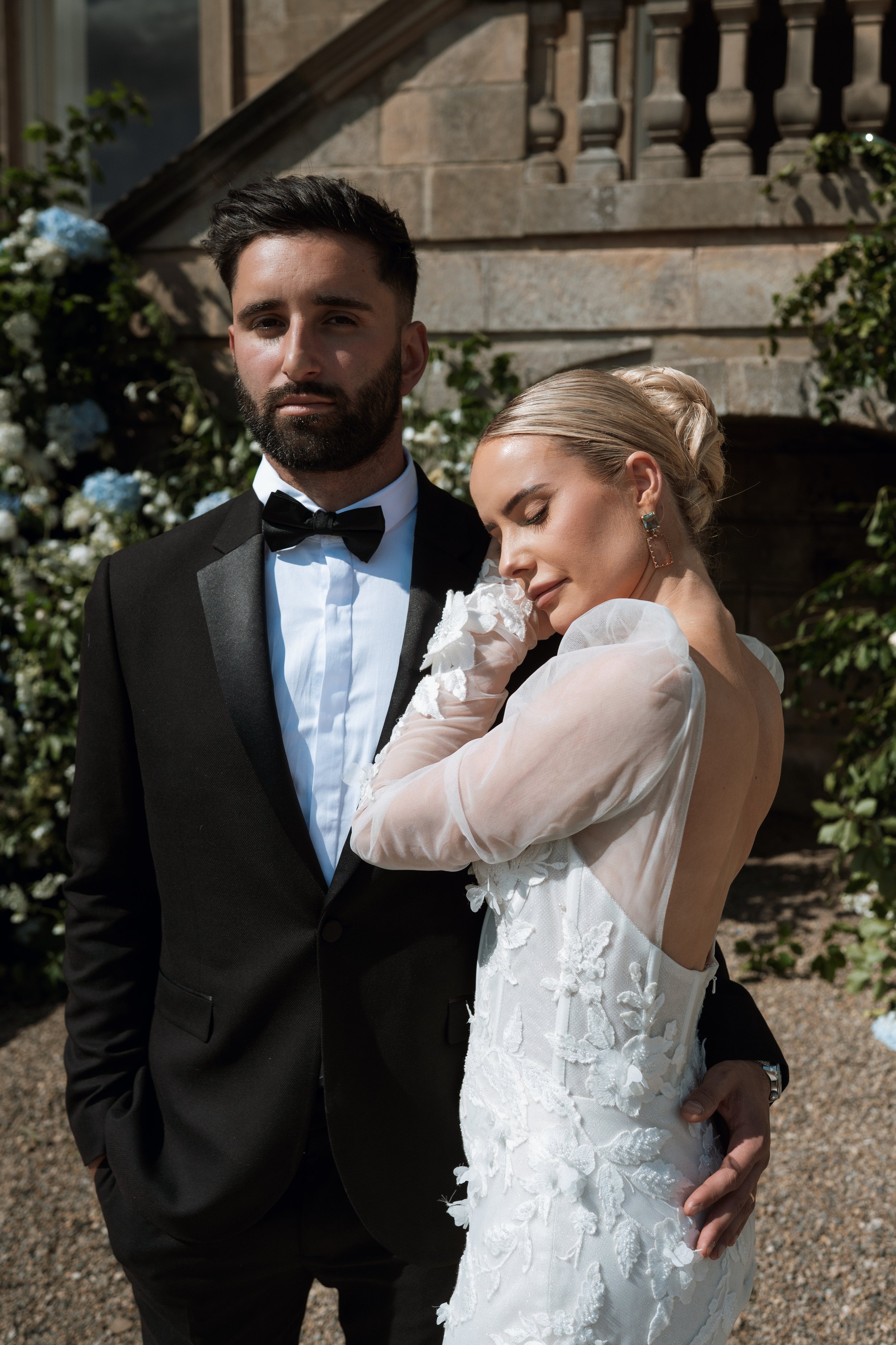 A bride and groom in wedding attire outdoors, with the bride in a white lace dress and the groom in a black tuxedo, standing in front of flowering bushes and a stone building.