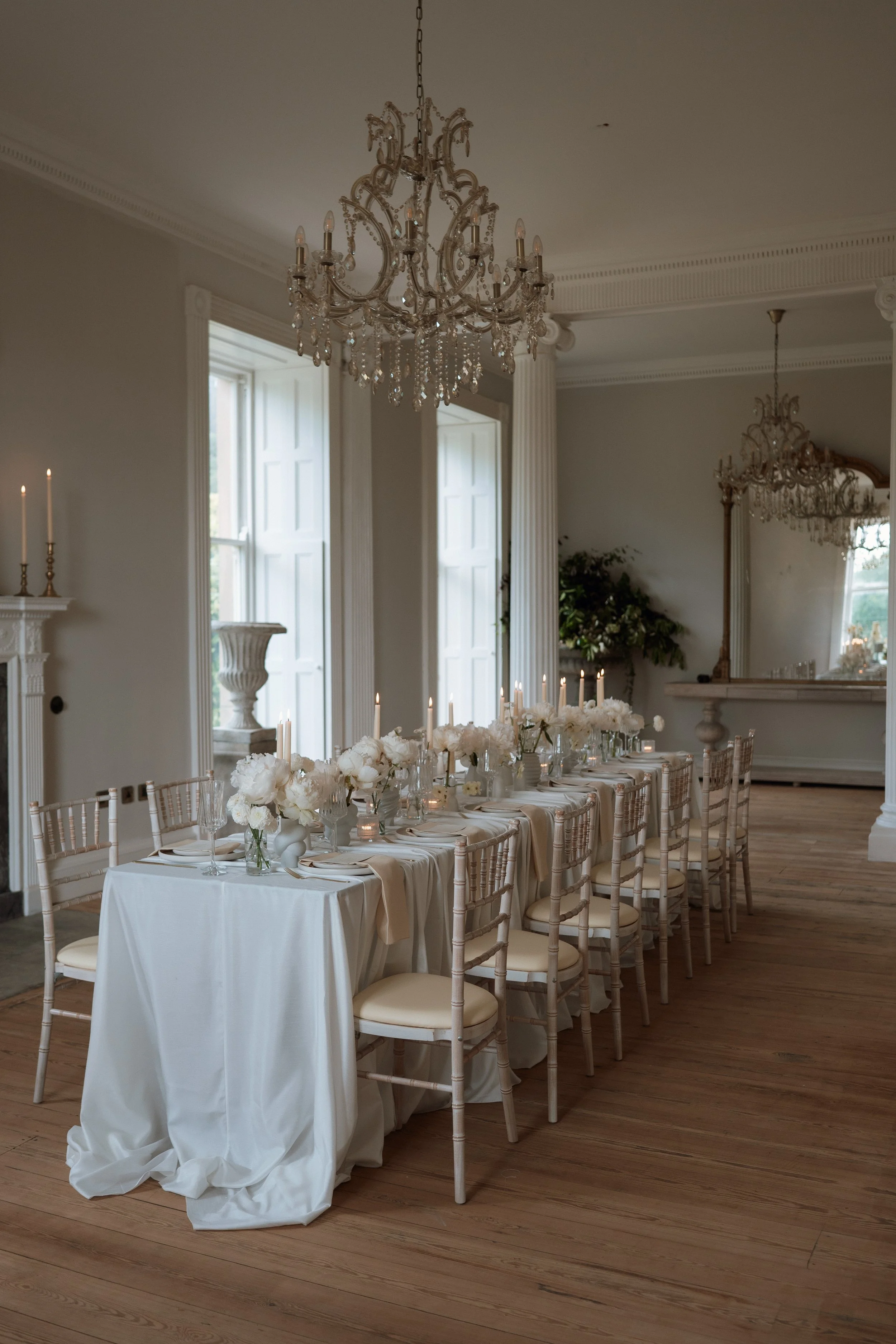 Elegant dinner table setup in a bright, decorated room with chandeliers, white flowers, candles, and classic chairs.